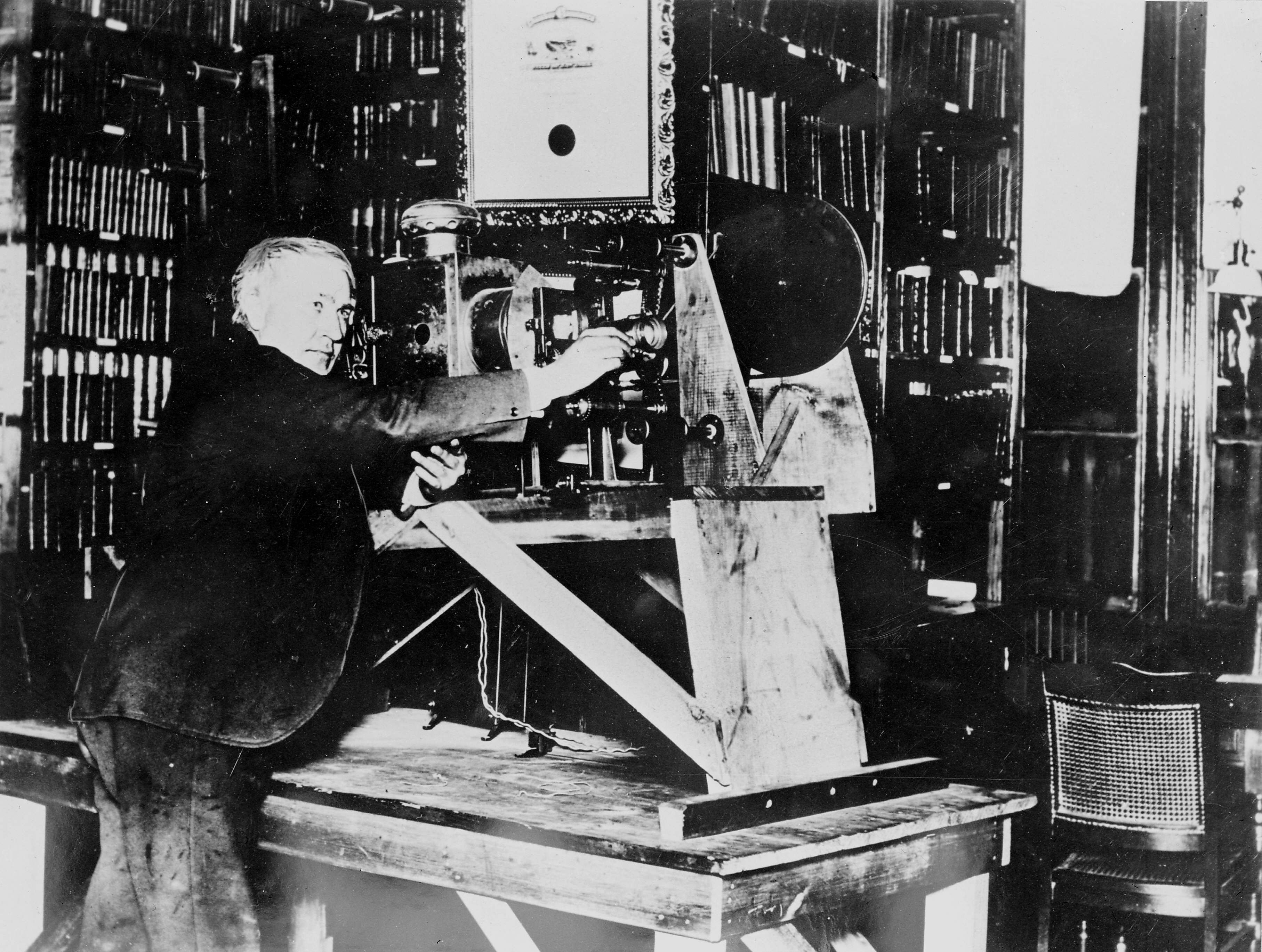 Black and white photo of an older man adjusting a large, vintage film camera on a wooden table in a room filled with bookshelves and chairs.