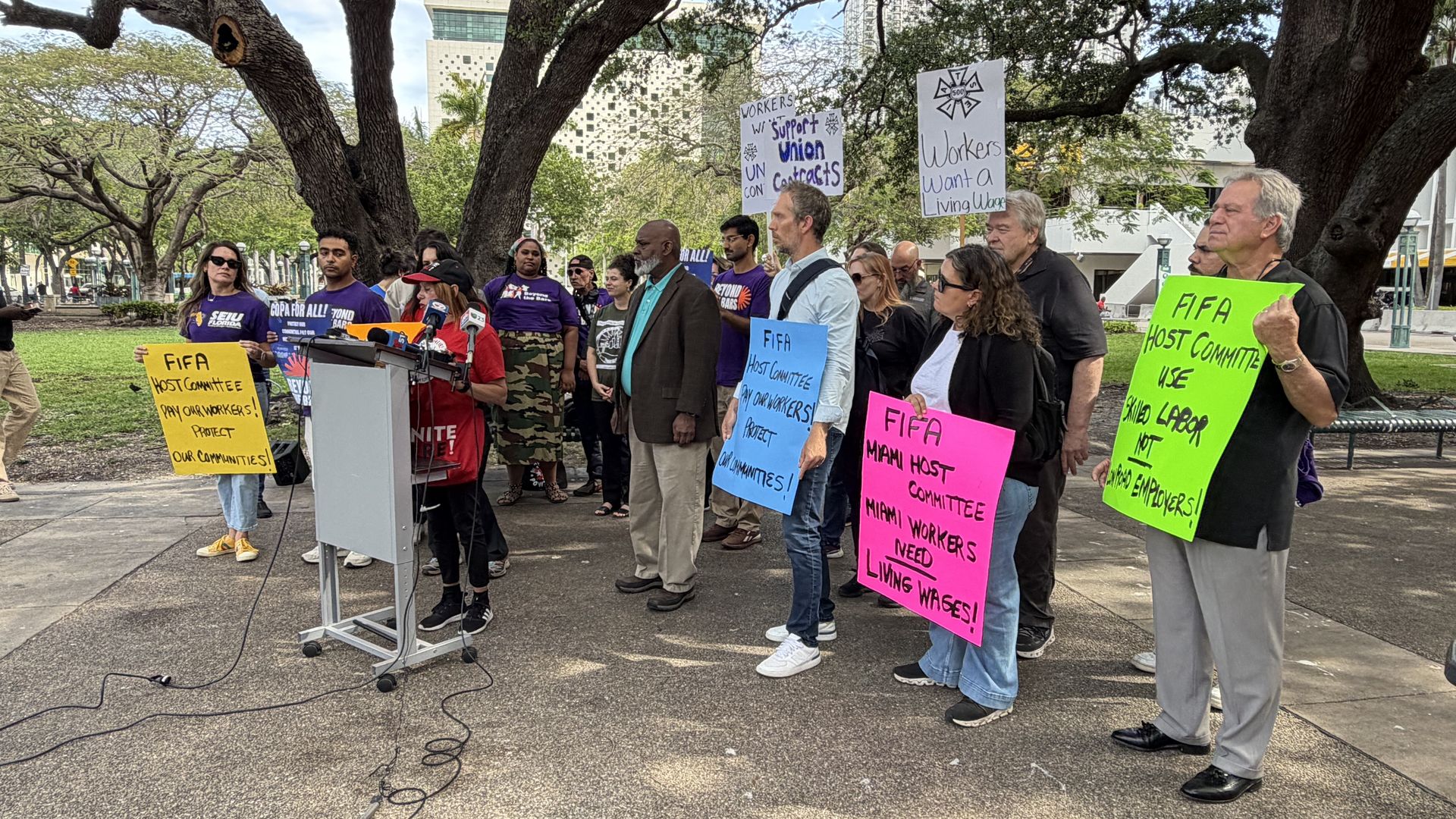Protesters in a park under large trees hold bright signs about FIFA workers' rights and living wages as a speaker addresses them at a podium with microphones on the ground.