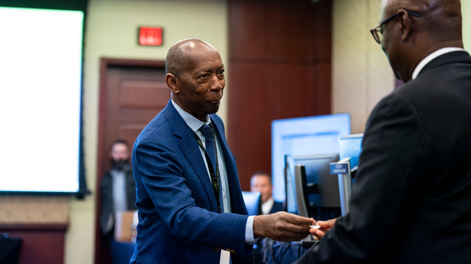 Rep. Sylvester Turner wearing a blue suit and reaching out his hand to a congressional staffer in a white and brown wooden room.