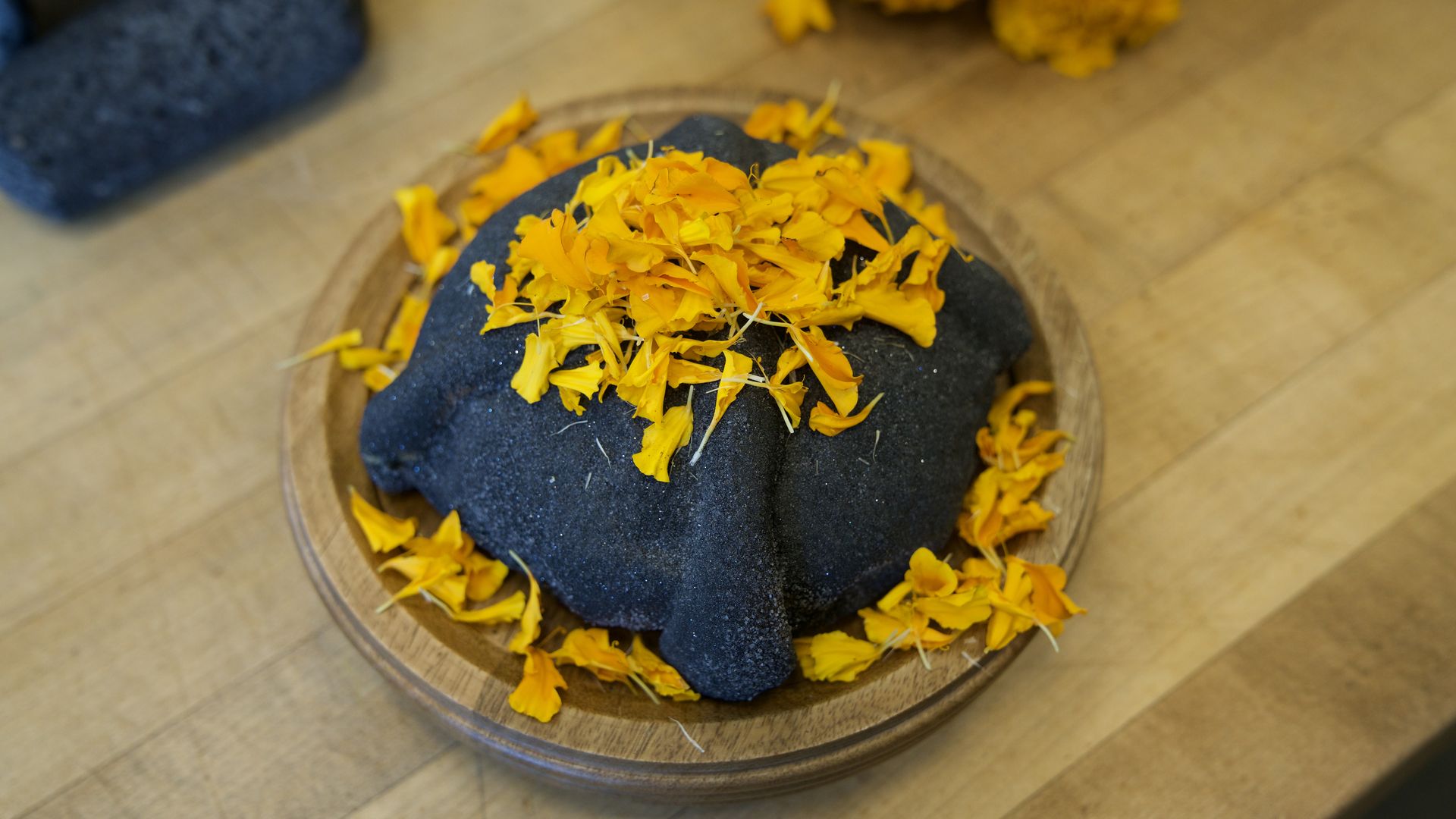 A circular black bread on a round wooden plate, decorated with scattered bright orange flower petals. The black bread has a shimmer to it.