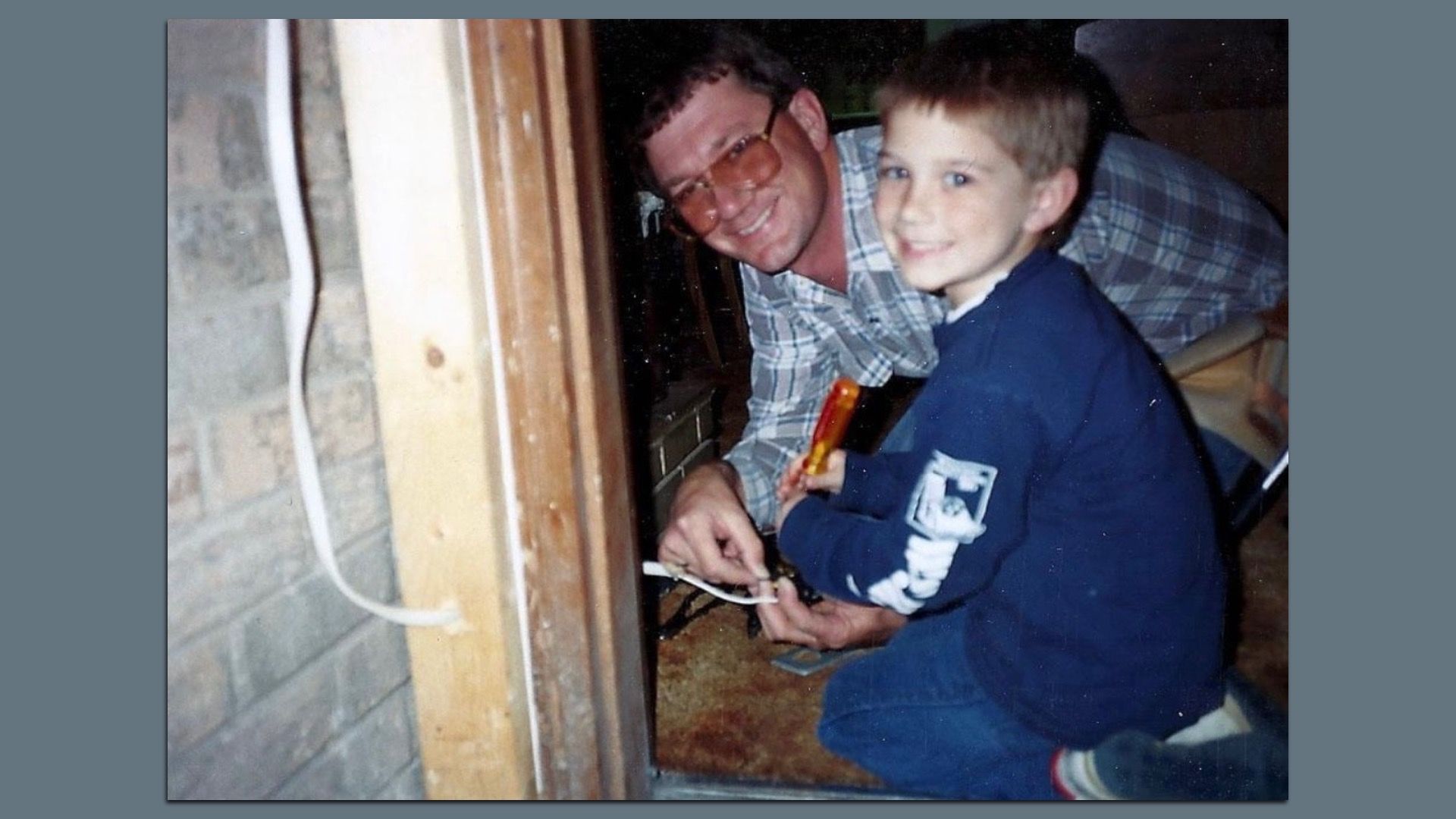 A man in a plaid shirt and orange-tinted glasses kneels beside a smiling boy in a blue hoodie, both looking at the camera while working near a doorway with a wooden frame and brick wall.