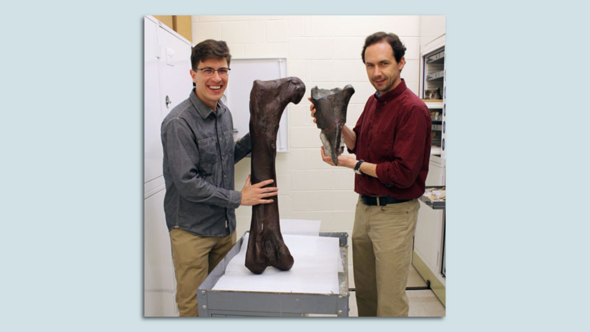 Two men stand side by side, one holding a large bone and another a fragment of a bone.