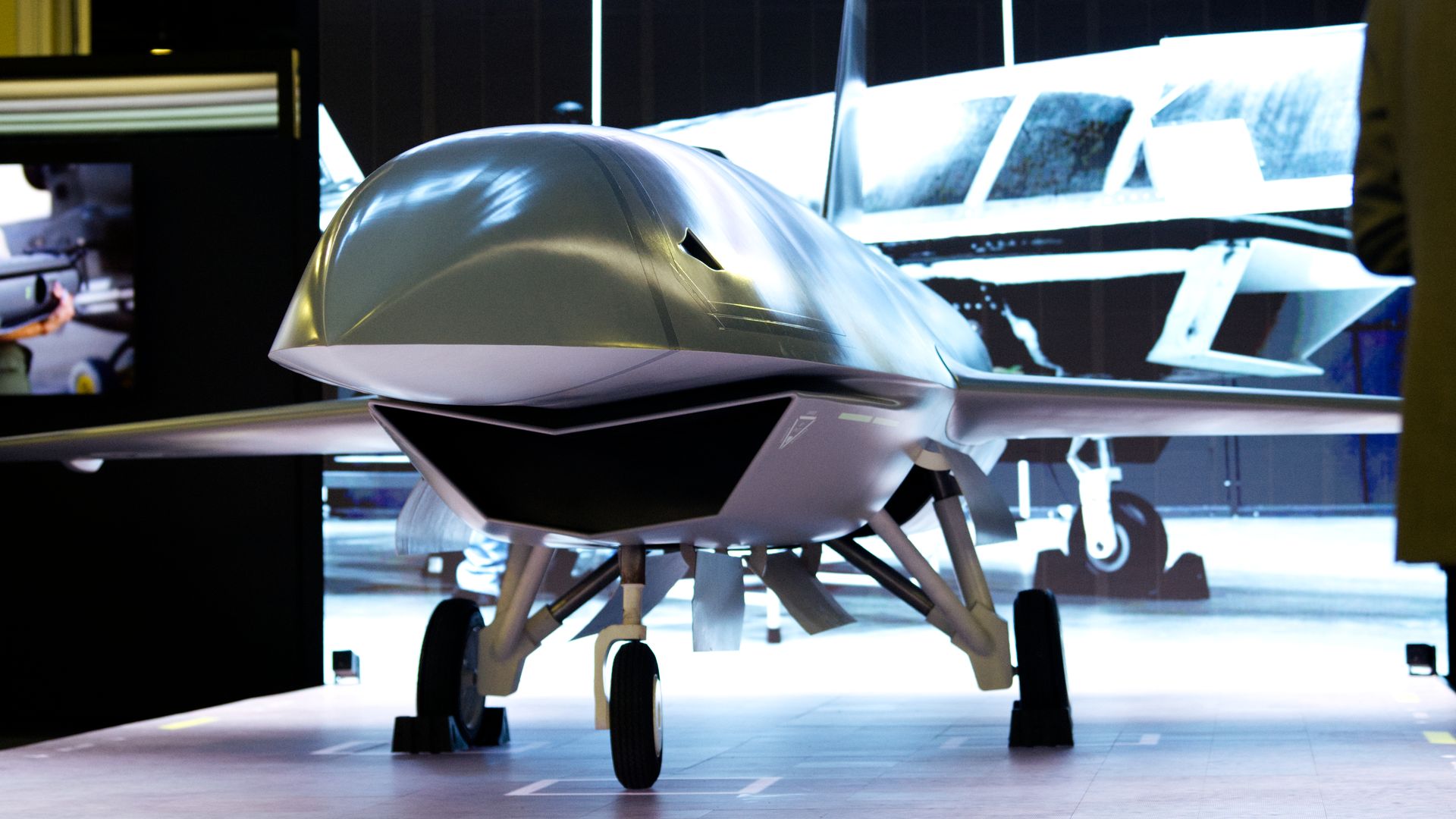 Front view of a sleek, silver unmanned aerial vehicle (UAV) with landing gear, displayed indoors with a digital screen background showing a similar drone blueprint.