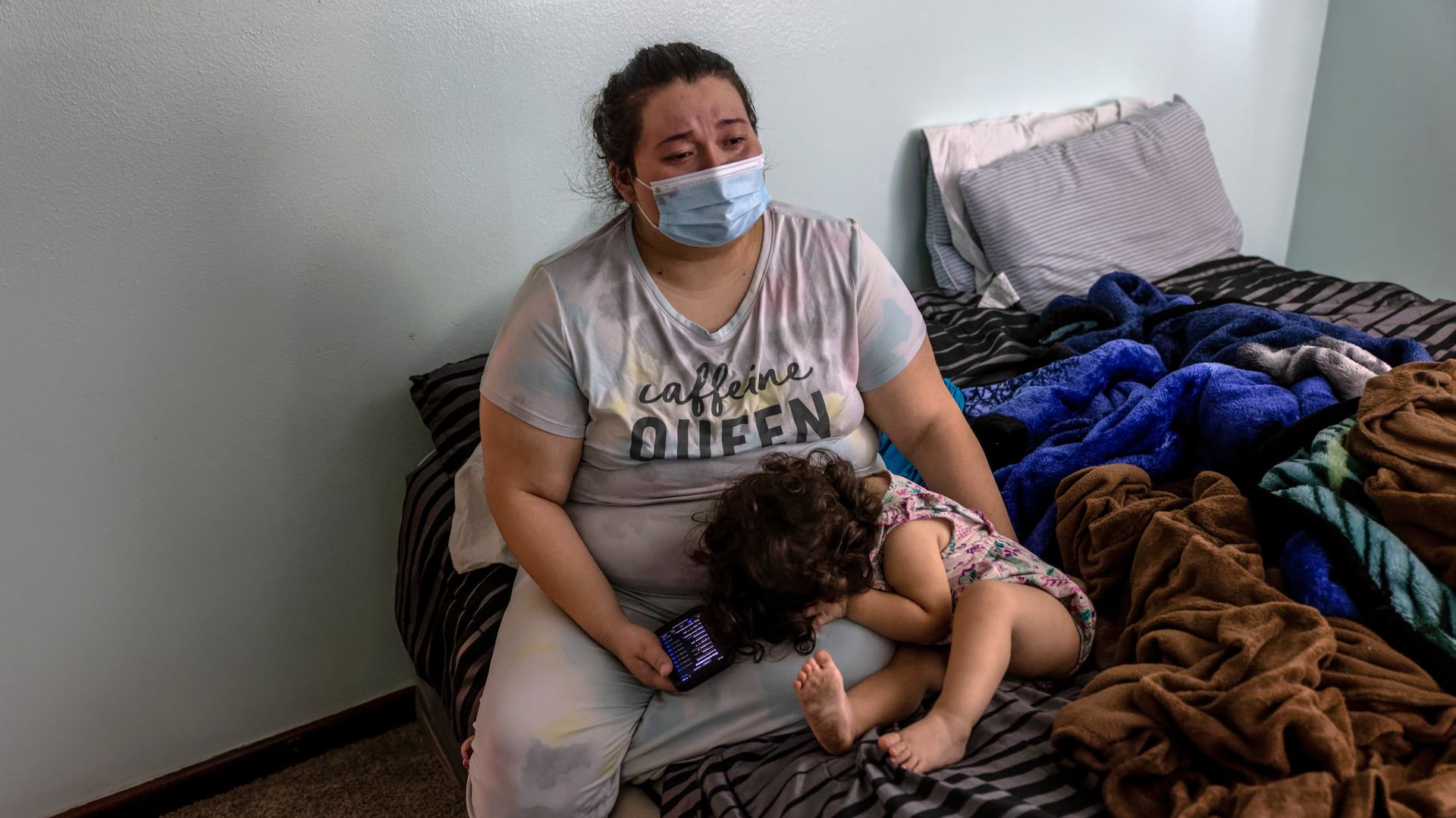 Picture of a woman wearing a mask sitting on the edge of a bed holding a child