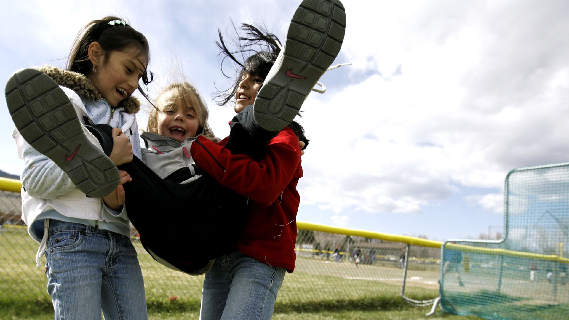Three children outdoors near a yellow fence and sports field; two girls in jeans and jackets lift a smiling child in gray and black jacket, sky cloudy overhead.