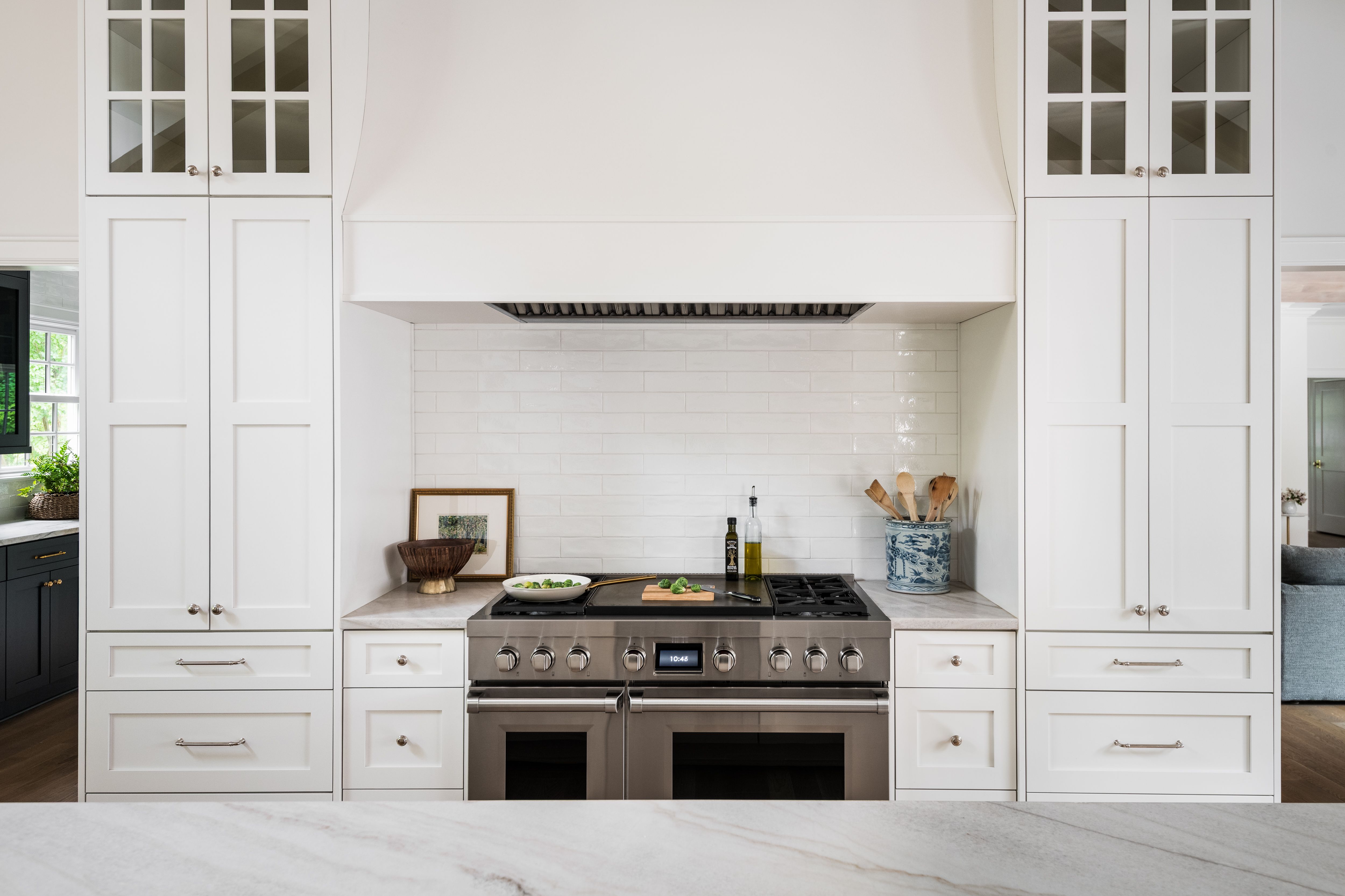 Modern white kitchen with stainless steel stove centered between tall white cabinets, a marble countertop, olive oil bottles, wooden utensils in a blue vase, and a bowl with green vegetables on the stove.