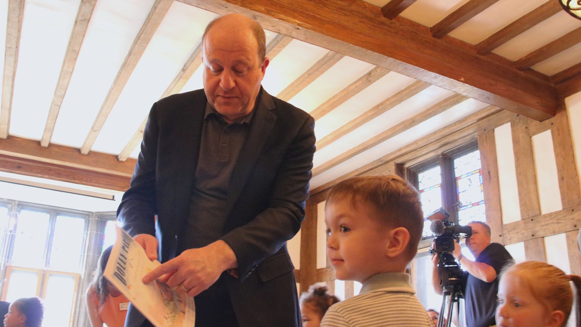 A man in a dark suit hands a map to a young boy in a striped shirt inside a wood-beamed room, with other children and a cameraman in the background.