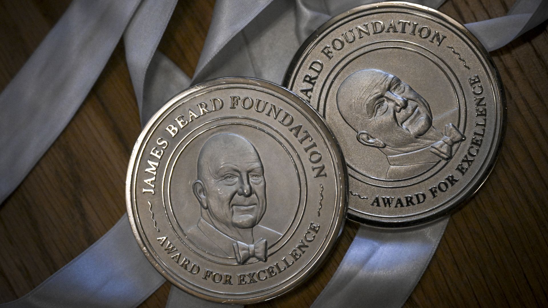 Two silver James Beard Foundation Award for Excellence medals on light gray ribbons, placed on a wooden surface.