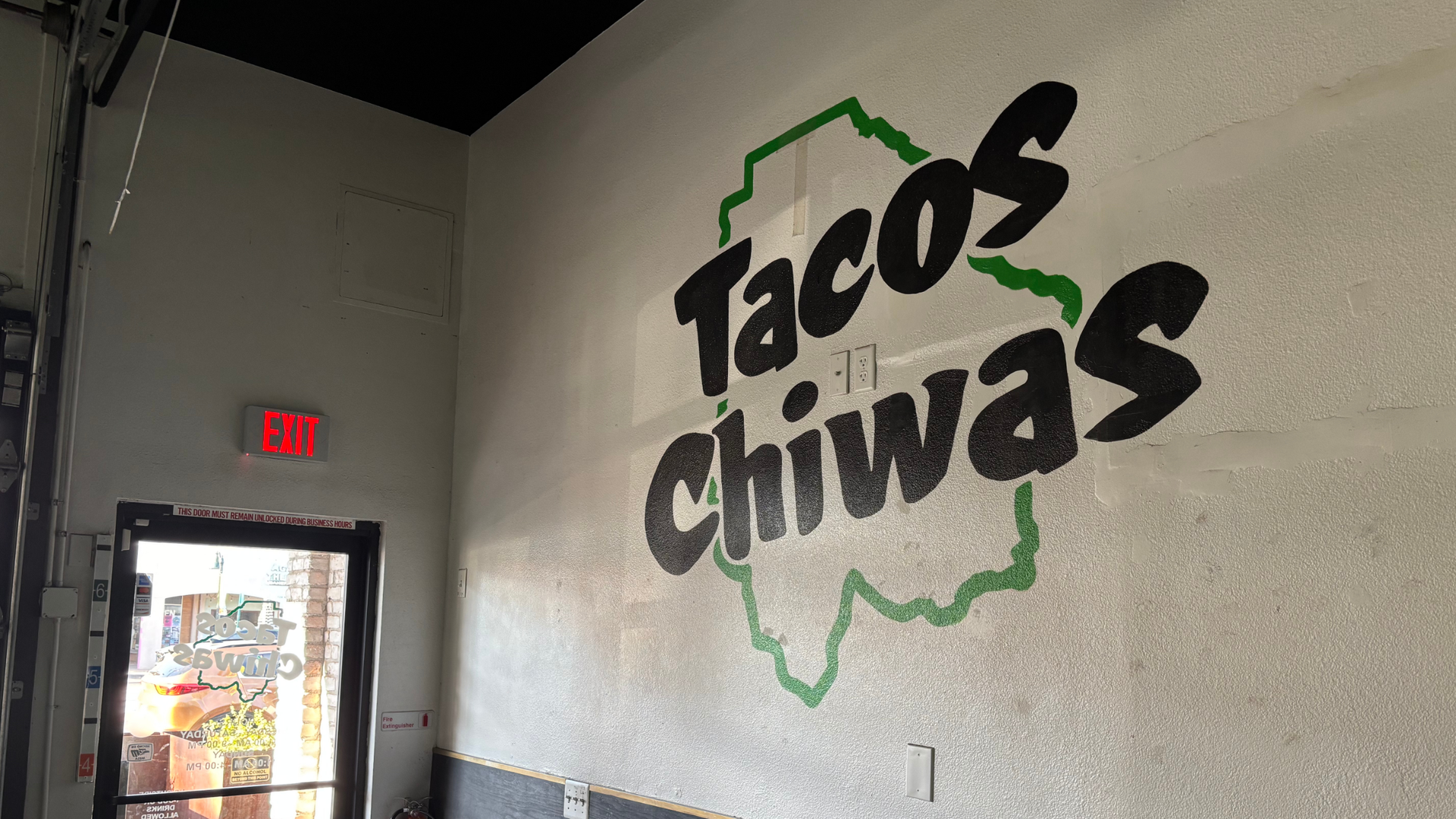 Indoor cafe with gray walls and a large black cursive mural accented in green on the right wall. A red EXIT sign sits above a glass door; wooden tables run along the lower wall.