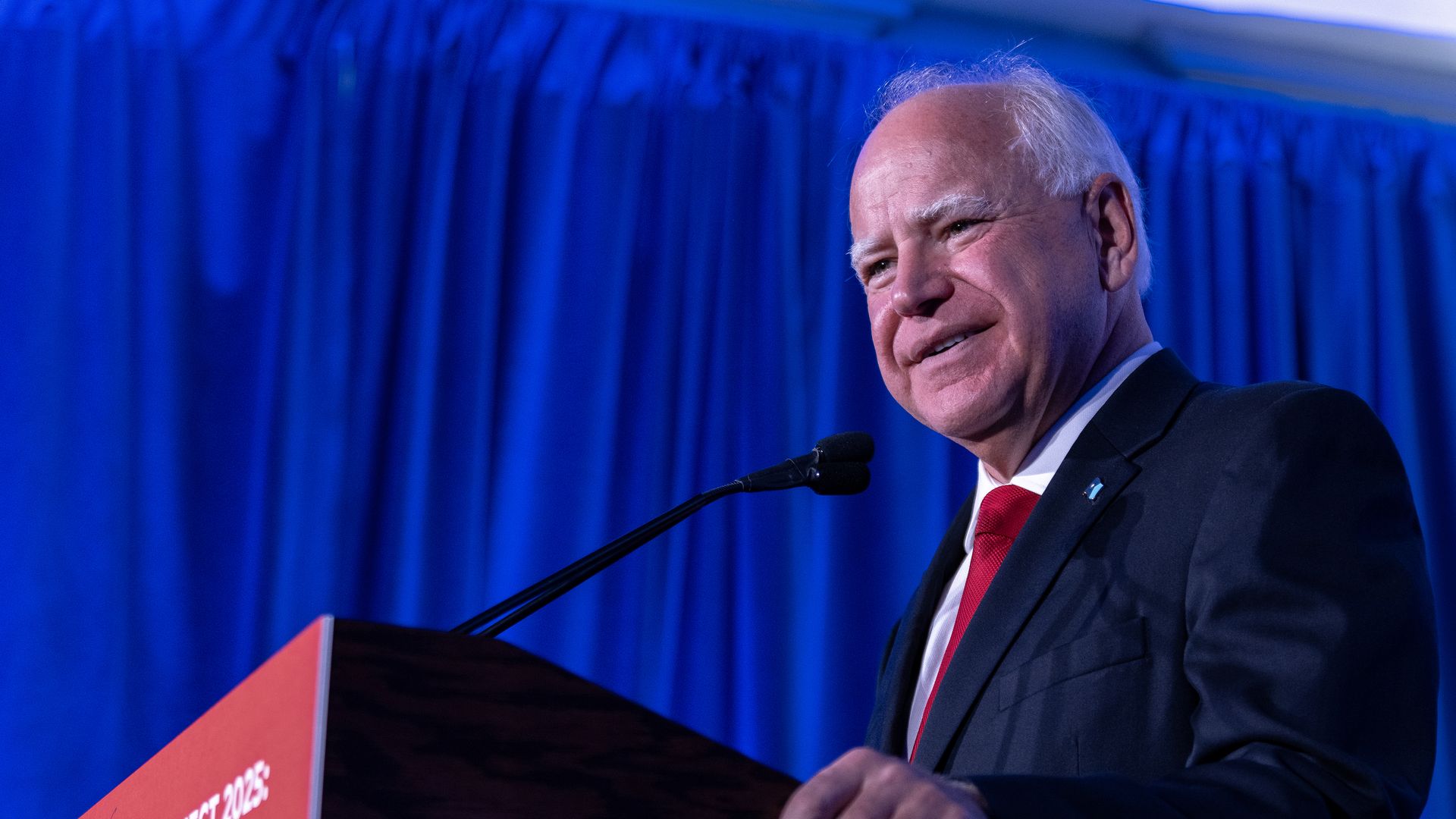 A man in a black suit, white shirt, and red tie speaks at a podium with a sign reading "Trump's Project 2025: Abortion, Punish Women" against a blue curtain backdrop.