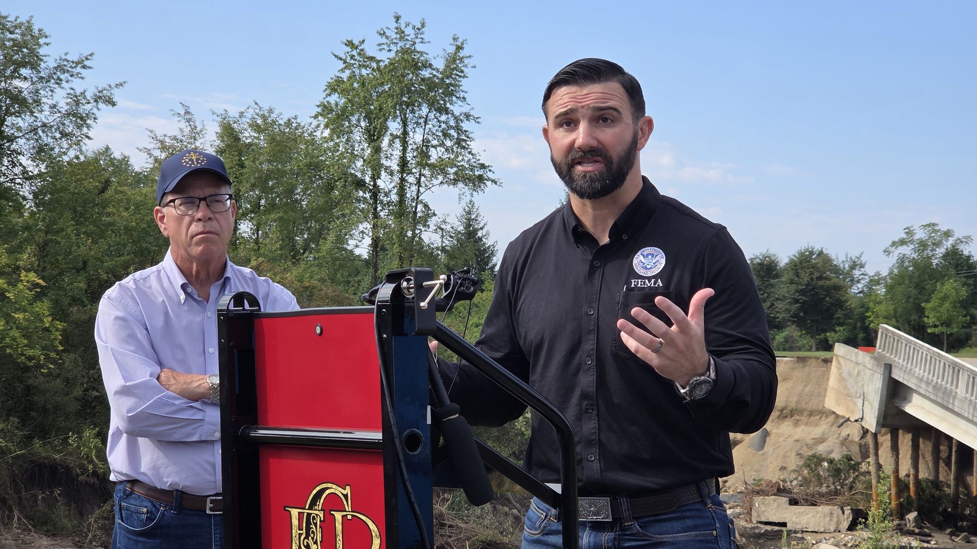 Two men stand outdoors beside a red and blue machine: one in a light shirt with a cap, the other in a black FEMA polo speaking, with trees, a blue sky, and a bridge in the background.