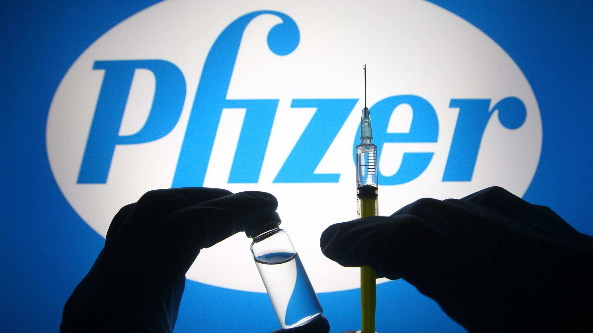 silhouette of hands in medical gloves hold a medical syringe and a vial in front of Pfizer logo