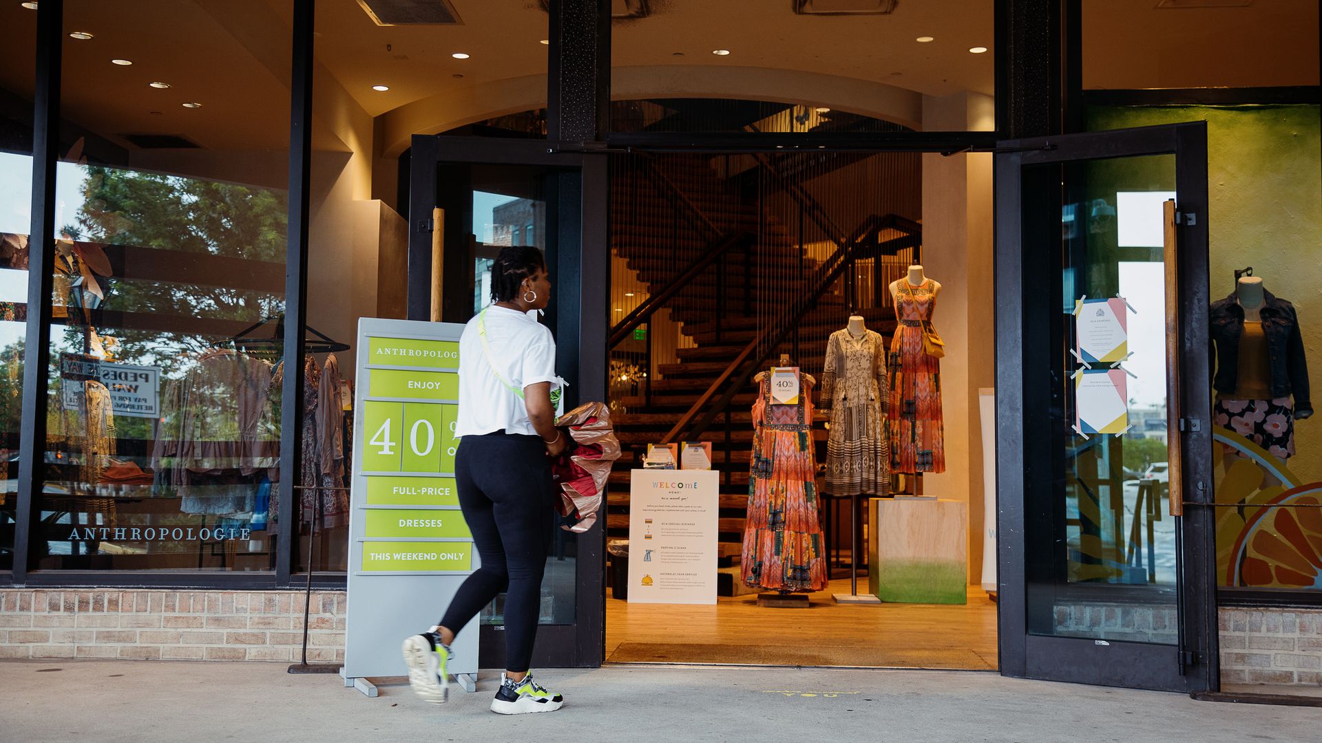 A woman walks into an Anthropologie store, which has its doors propped open. She walks past a sign displaying an ongoing sale.