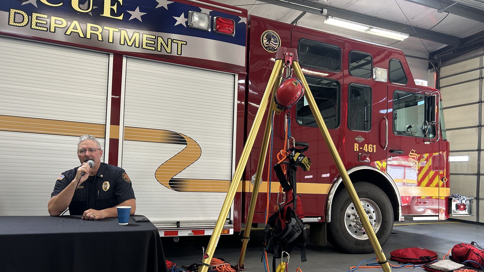 Firefighter speaks into a microphone at a table inside a fire station, with a large red fire engine behind him and tangled yellow hoses and gear scattered on the floor.
