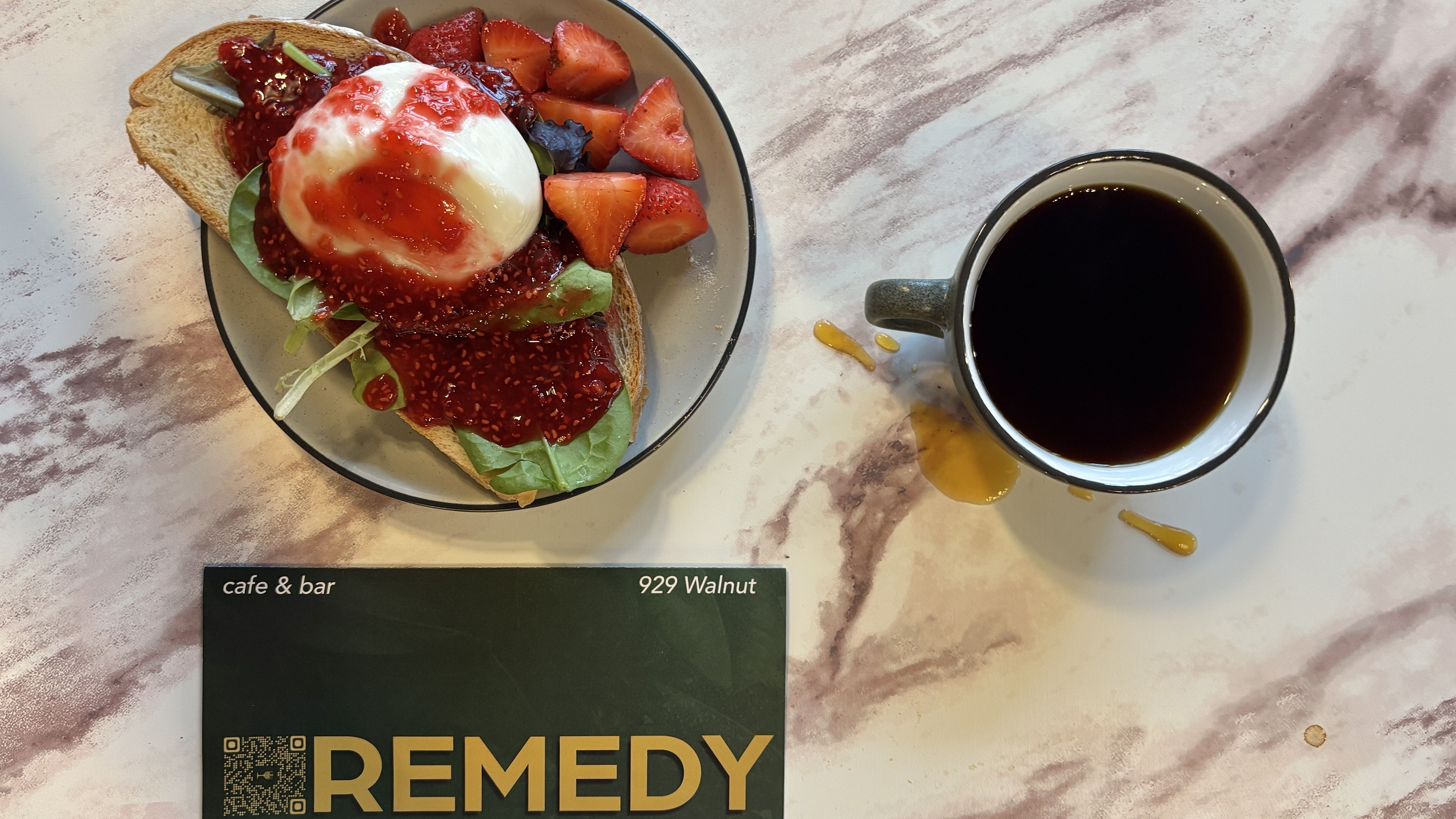Top-down view of toast with greens, a poached egg, and strawberry jam; sliced strawberries on the side. A black coffee cup sits on a marble table, beside a cafe menu card reading REMEDY.