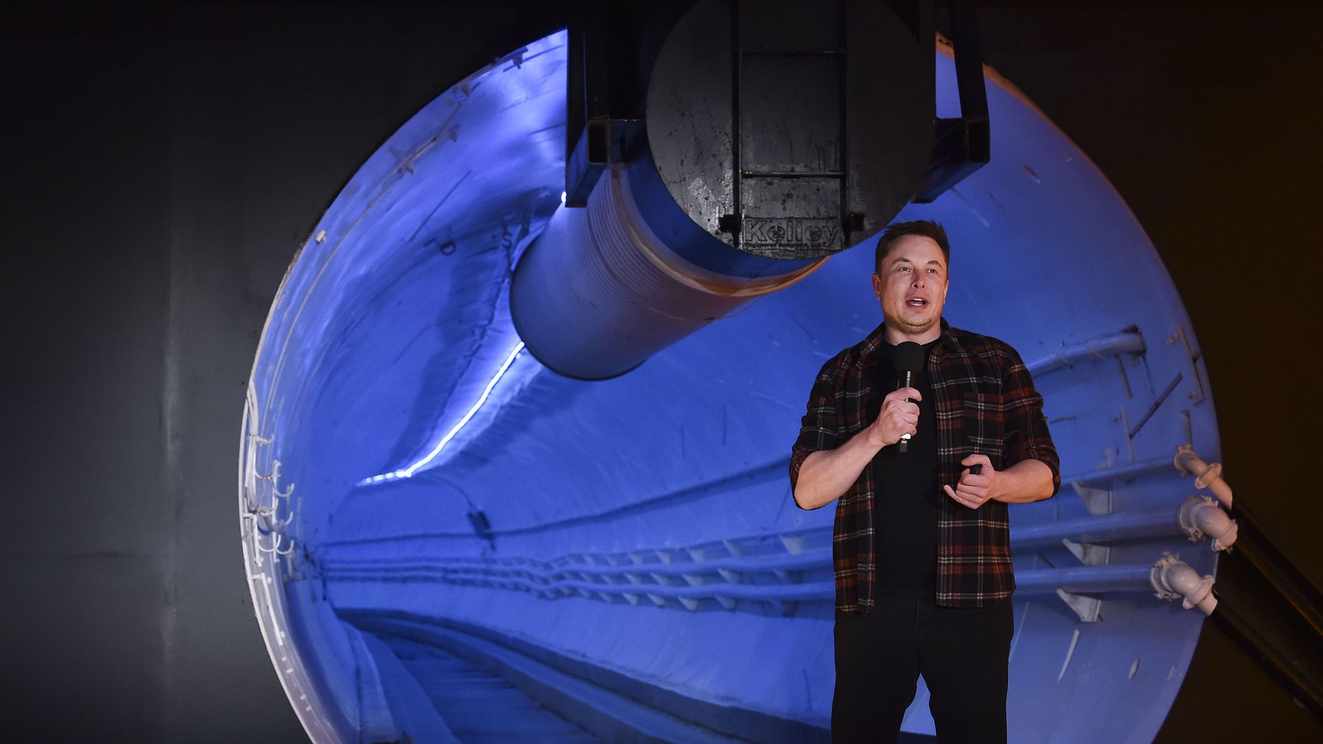 In this image, Elon Musk stands and holds a microphone in front of a tunnel opening.