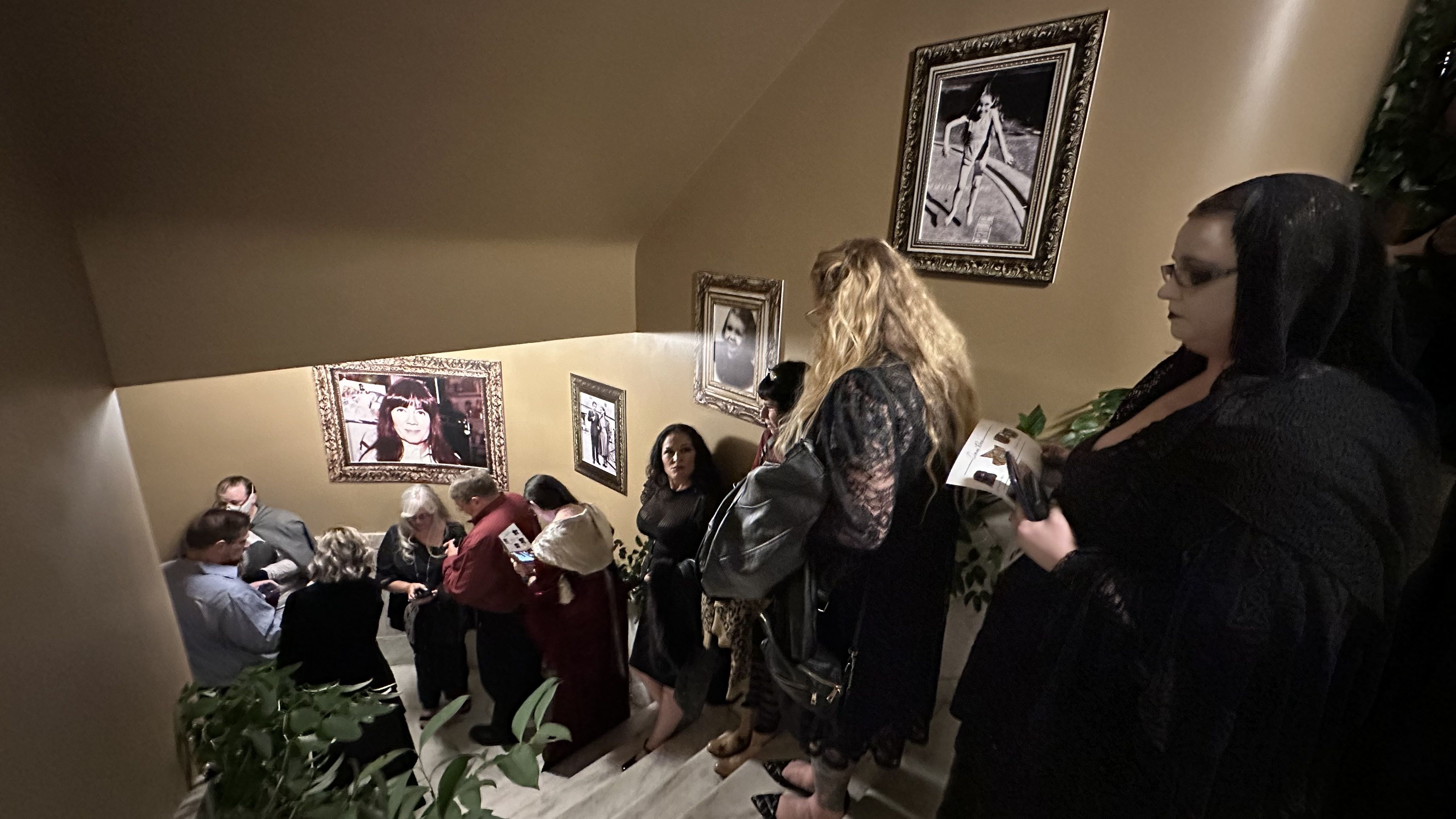 Group of people dressed in black and dark clothing standing on a beige stairway with framed black-and-white and color portraits on the walls.