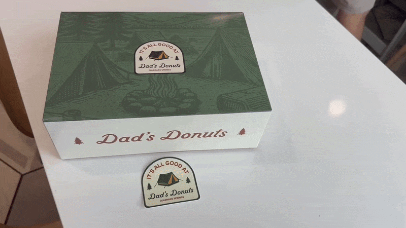 A hand opening a half-doxen box of Dad's Donuts. Inside the box are an assortment of donuts, including pink, chocolate and vanilla frosted. 