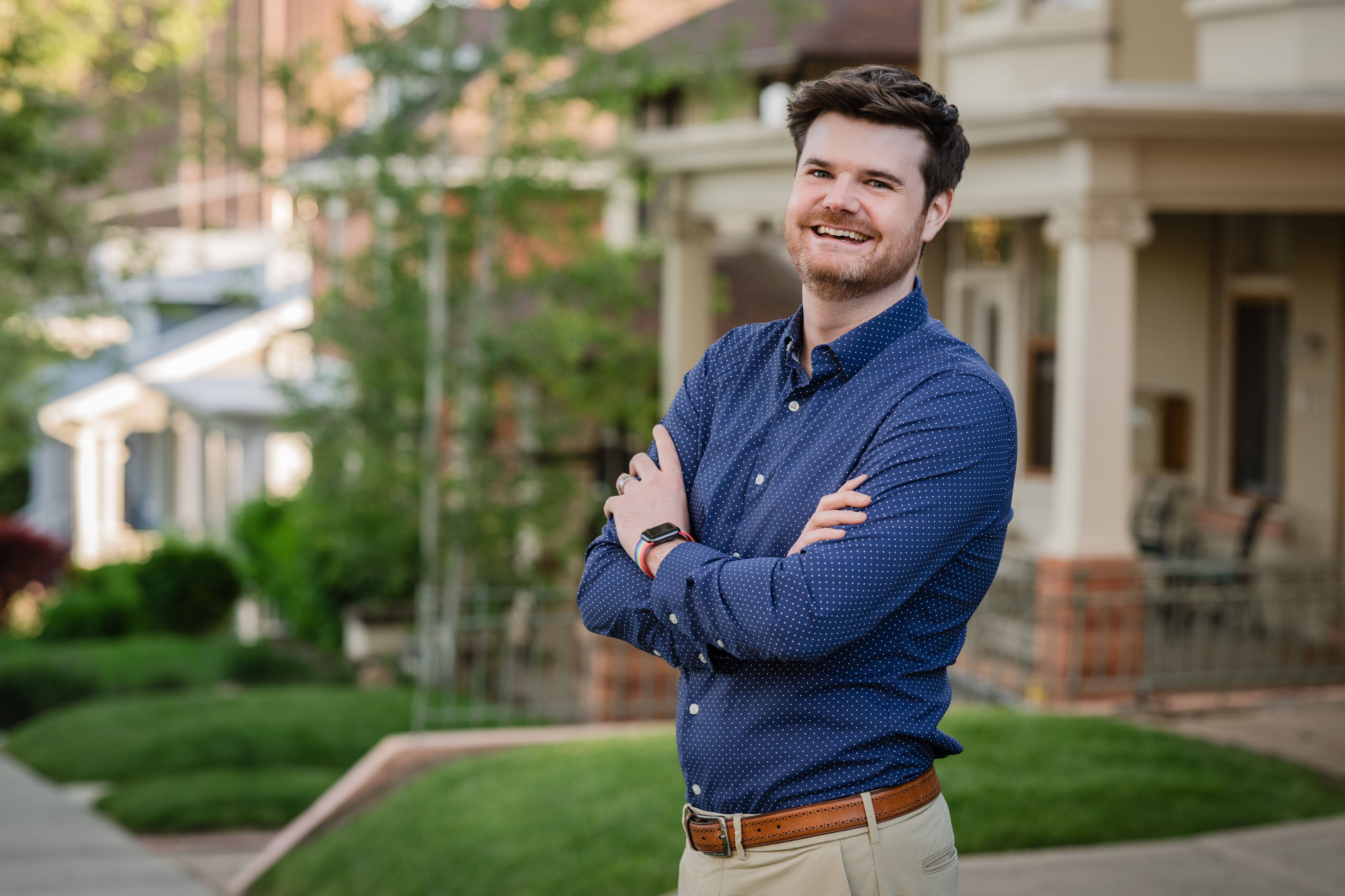 Smiling man with brown hair and beard wearing a blue polka-dot shirt and beige pants, standing outdoors with arms crossed in front of residential houses and greenery.