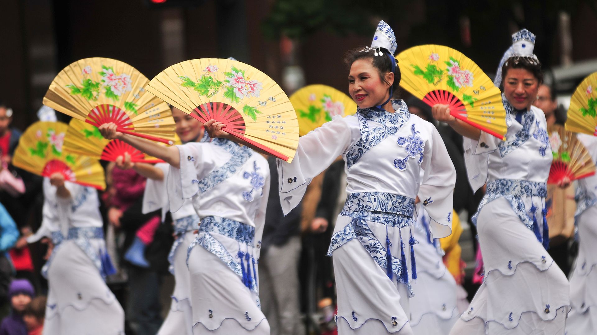 Women in traditional Chinese dresses perform a fan dance during a parade, wearing white and blue costumes and holding yellow fans decorated with flowers, as a crowd watches in the background.