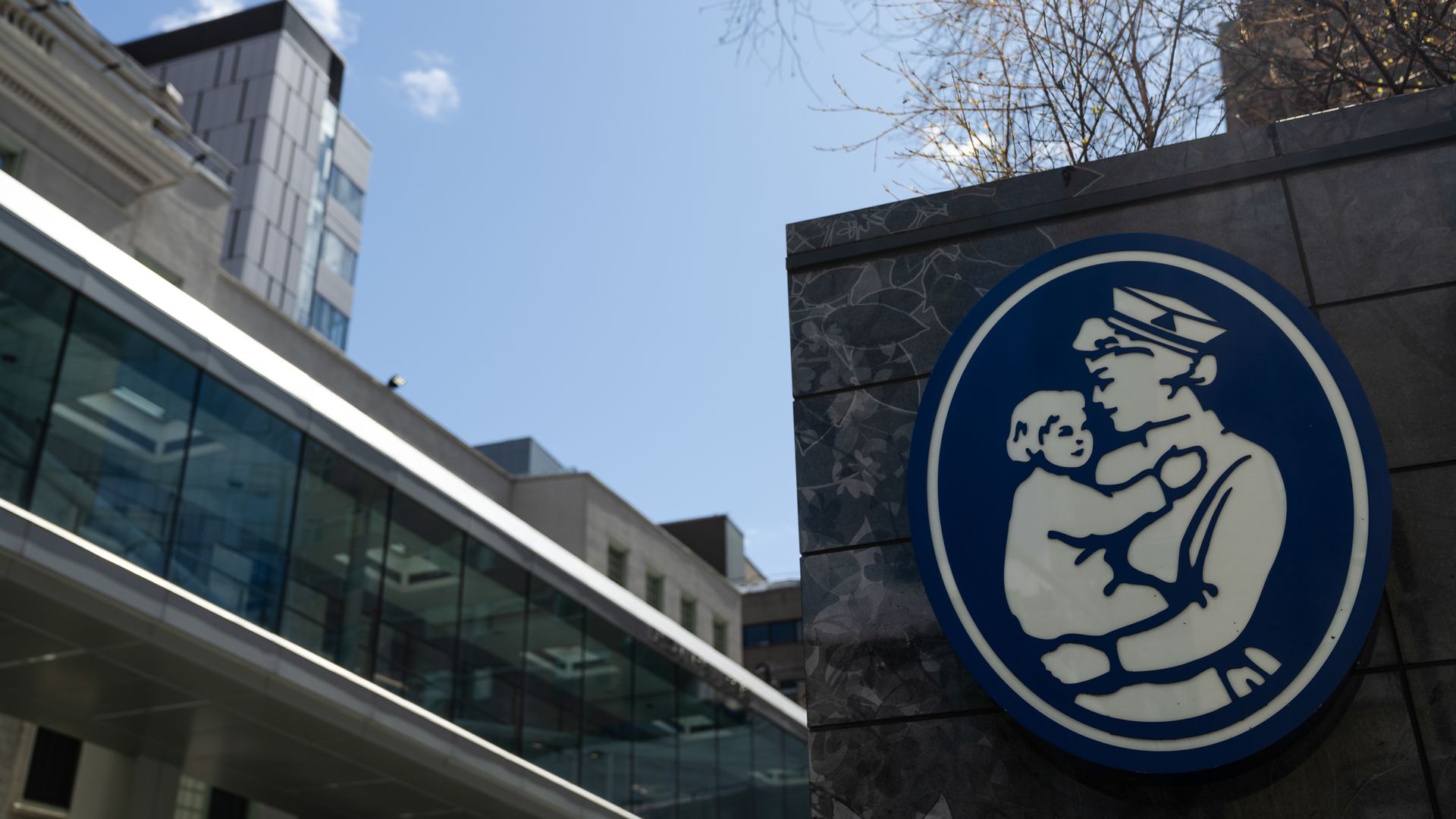 Blue and white circular sign showing a person in a uniform holding a child, mounted on a dark tiled wall with modern buildings and a blue sky in the background.