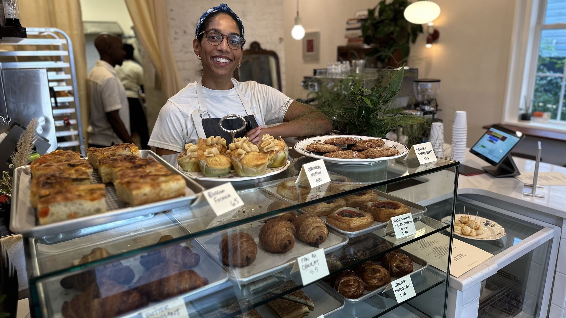 A woman sits behind a pastry case and smiles at the camera.