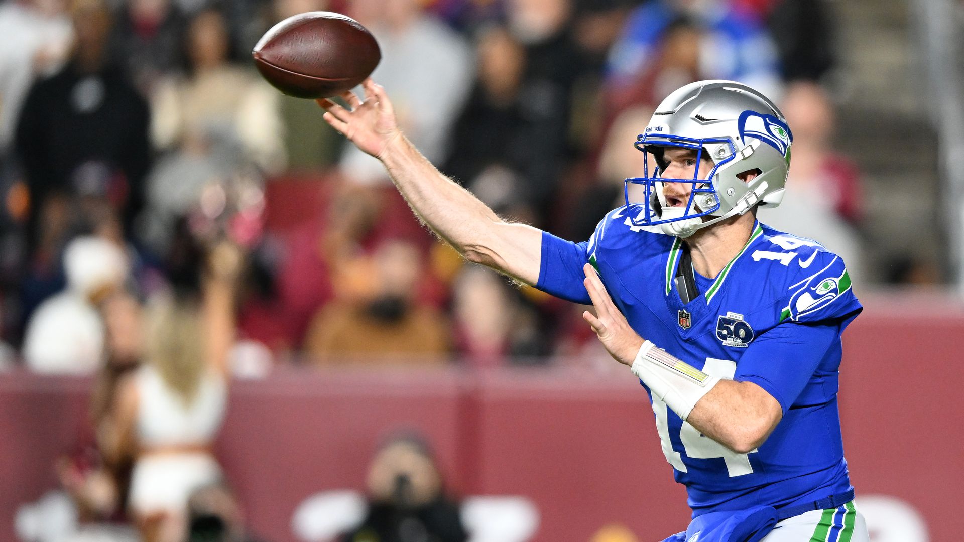 Sam Darnold in a blue jersey and silver helmet with Seahawks logo is shown with the ball just leaving his hand as he throws, with a blurry crowd in the stadium in the background.