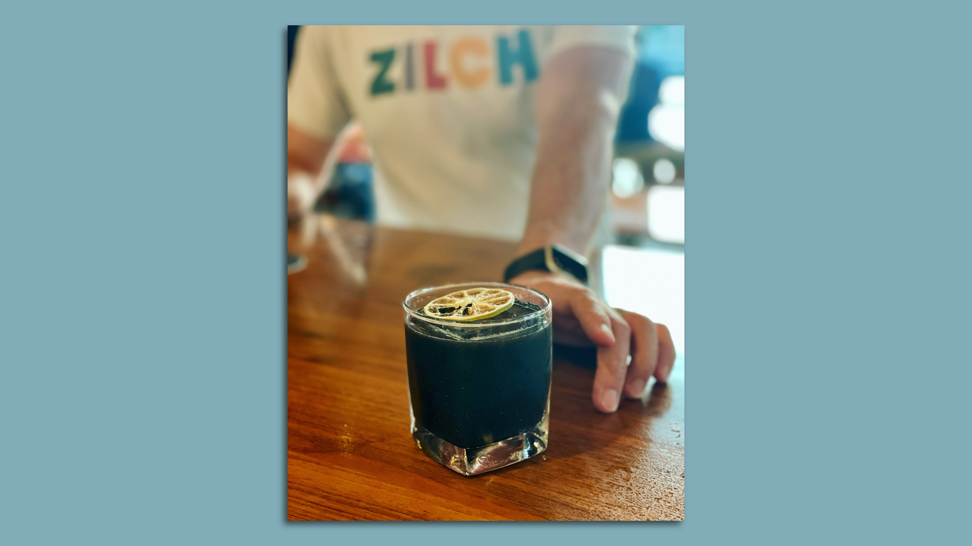 A person wearing a shirt that says "Zilch" slides a dark cocktail with a dried orange across a stained wood bar.
