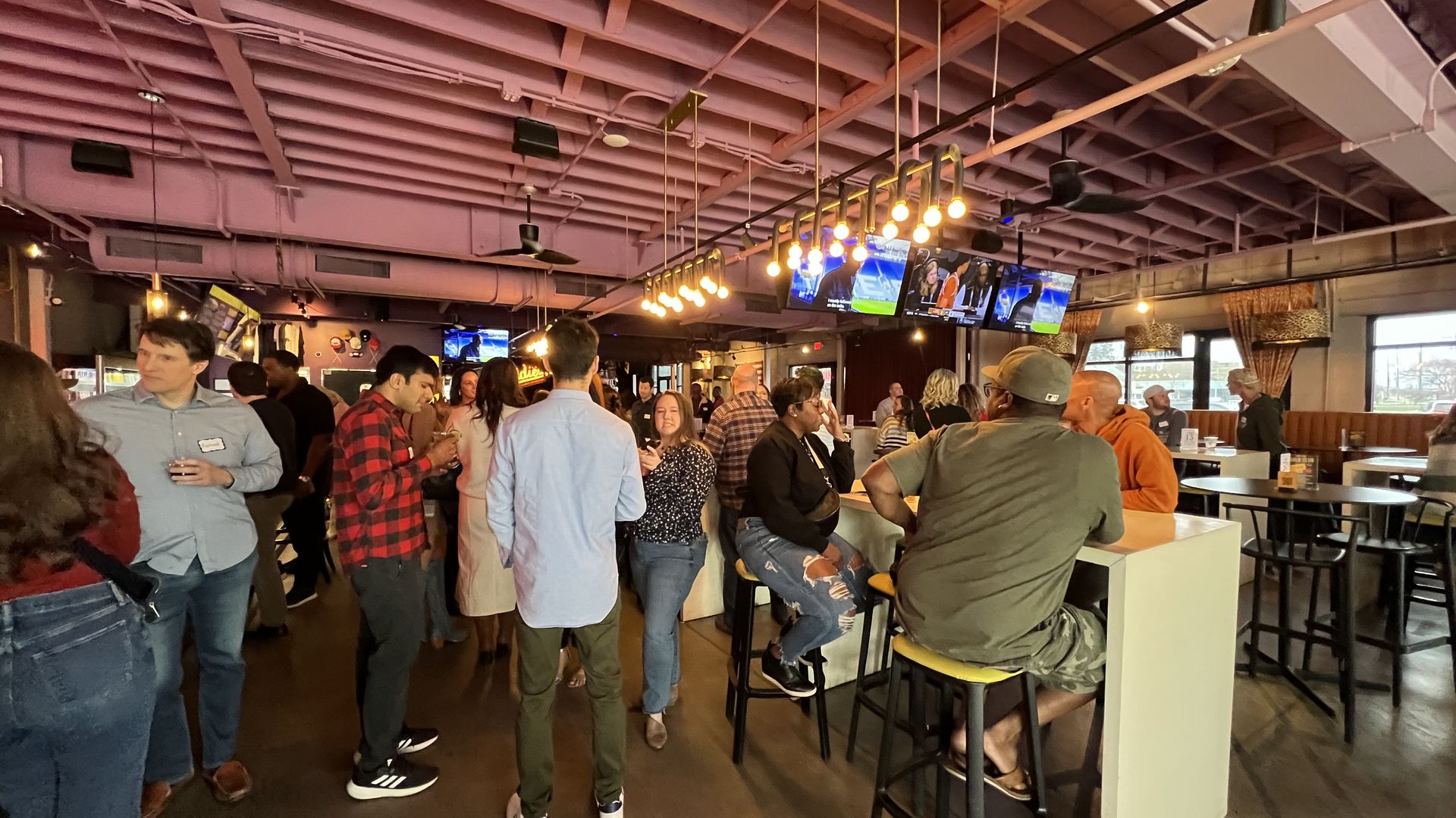 A crowded bar with people socializing and sitting on stools at high tables under warm hanging lights and purple-painted exposed beam ceiling, with TVs showing a sports game.