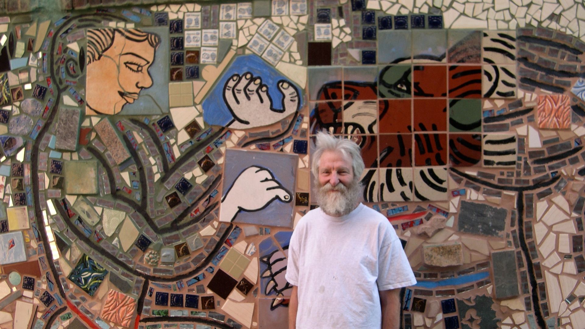 A bearded man in a white shirt and shorts stands smiling in front of a colorful mosaic wall depicting abstract shapes, hands, and a face using various tiles and patterns.