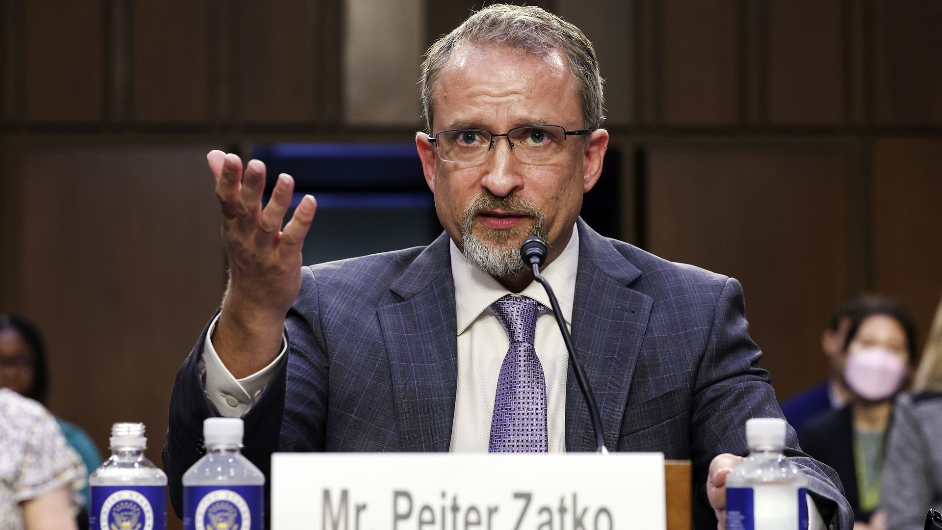 Photo of Peter Zaitko in a jacket and tie gesturing with a placard stating his name in front of him