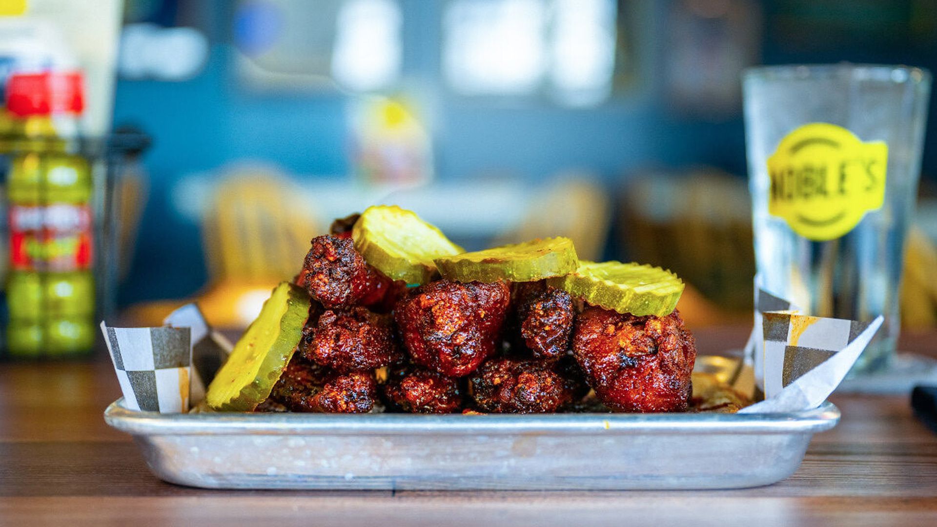 Tray of spicy, crispy fried chicken wings topped with several green pickle slices, set on a wooden table with a blurry blue background and a glass with a yellow logo nearby.