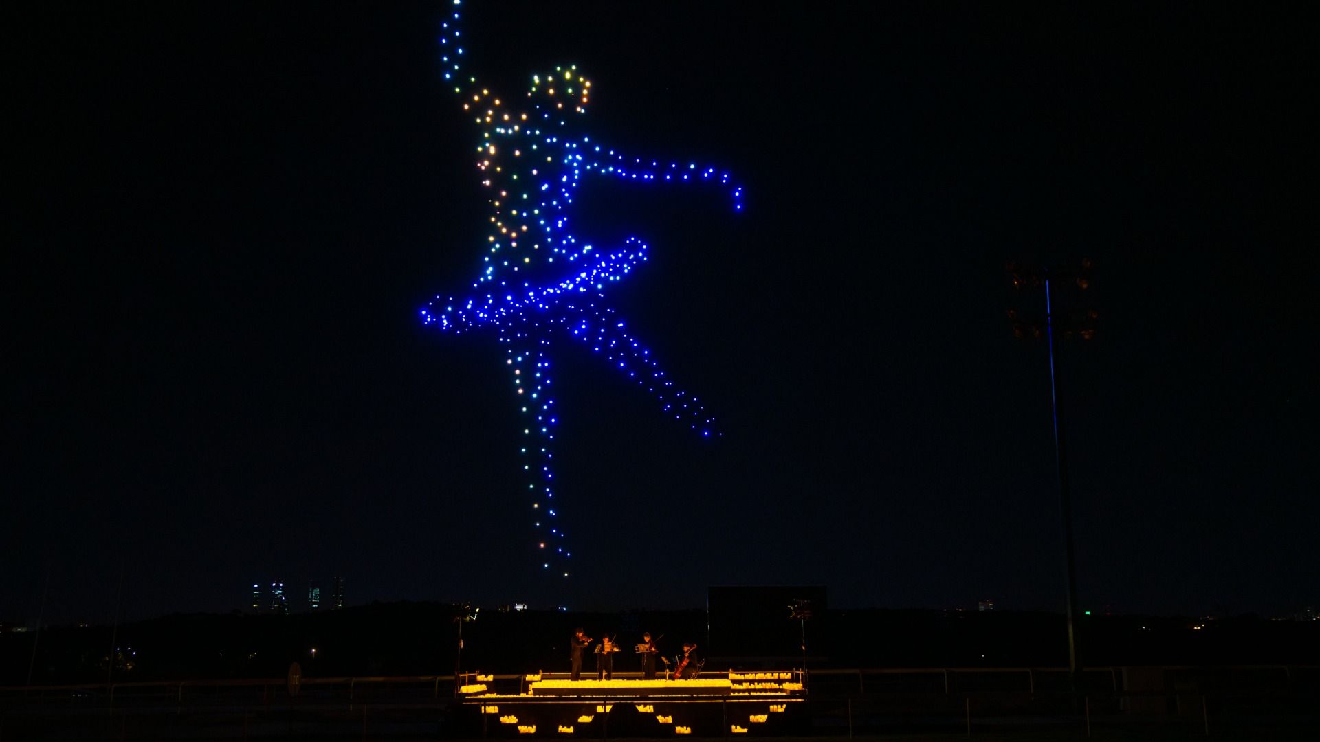 Night scene with a colorful drone light show forming a ballerina figure in blue, yellow, and orange lights above a small lit stage where musicians play string instruments.