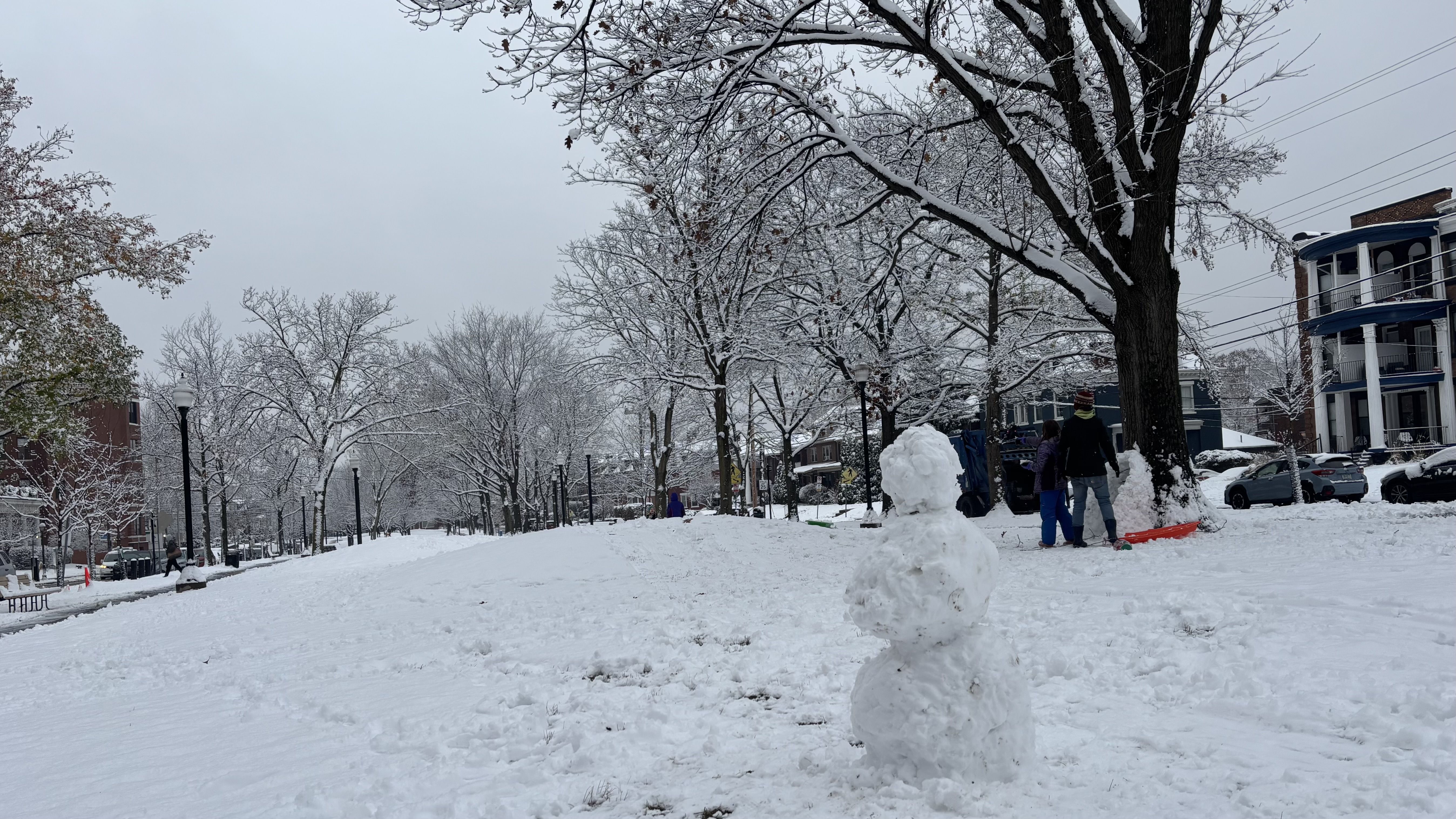 Snow-covered urban park with bare trees dusted in snow, a small snowman in foreground, people playing near large tree, and multi-story houses along the street on a cloudy winter day.