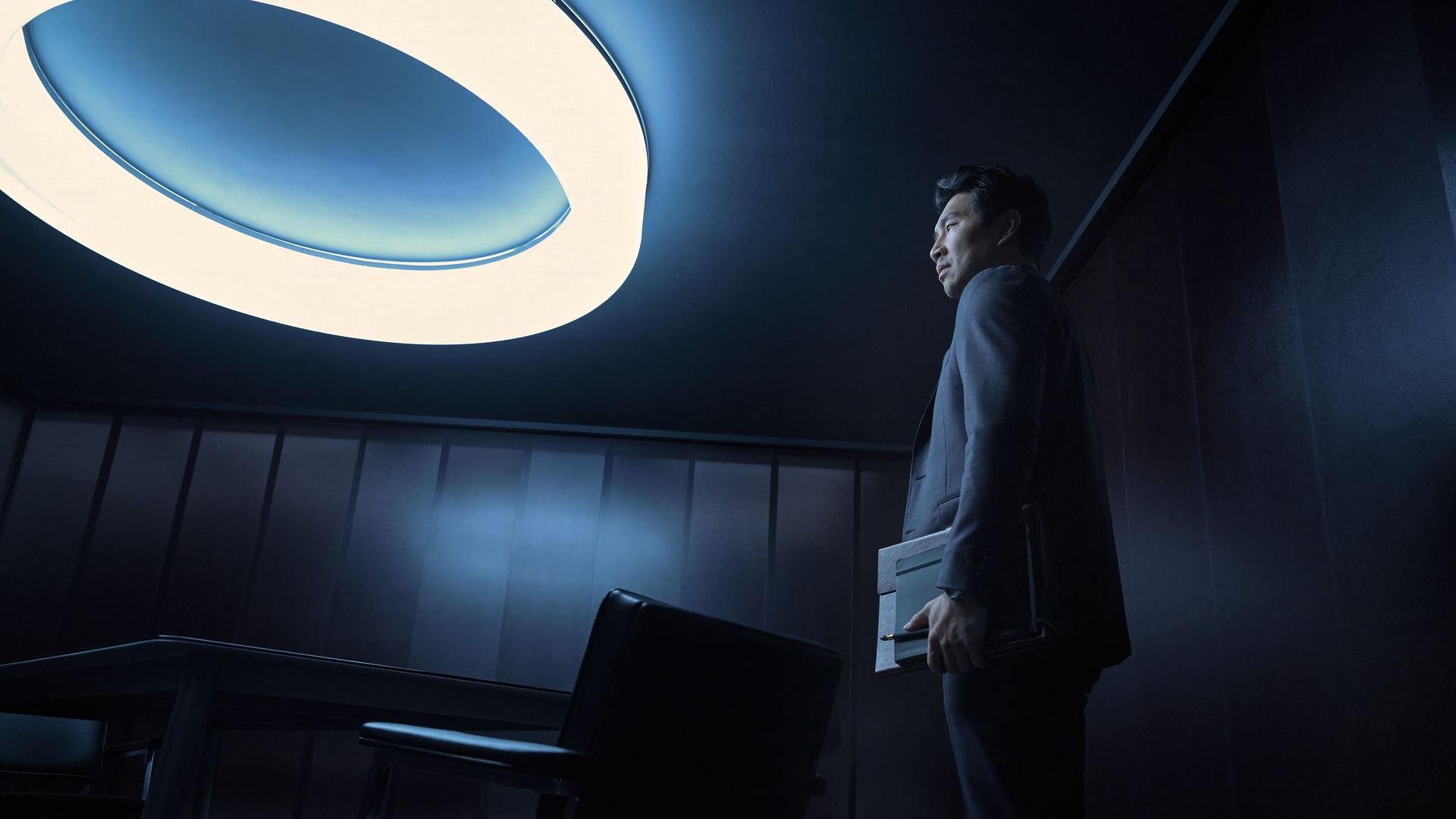 Man in a dark suit holding files, standing in a dimly lit room with dark walls, near a table and chairs, illuminated by a large circular ceiling light.