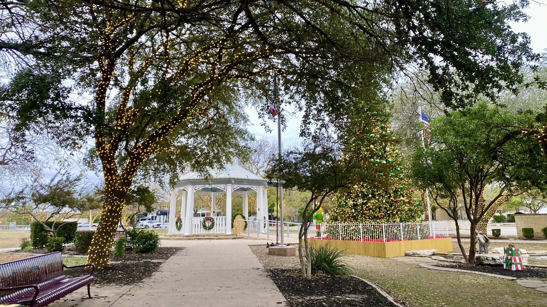 Park walkway with trees wrapped in white lights, a white gazebo decorated with green wreaths, and a large Christmas tree adorned with red, green, and gold ornaments surrounded by a white fence.