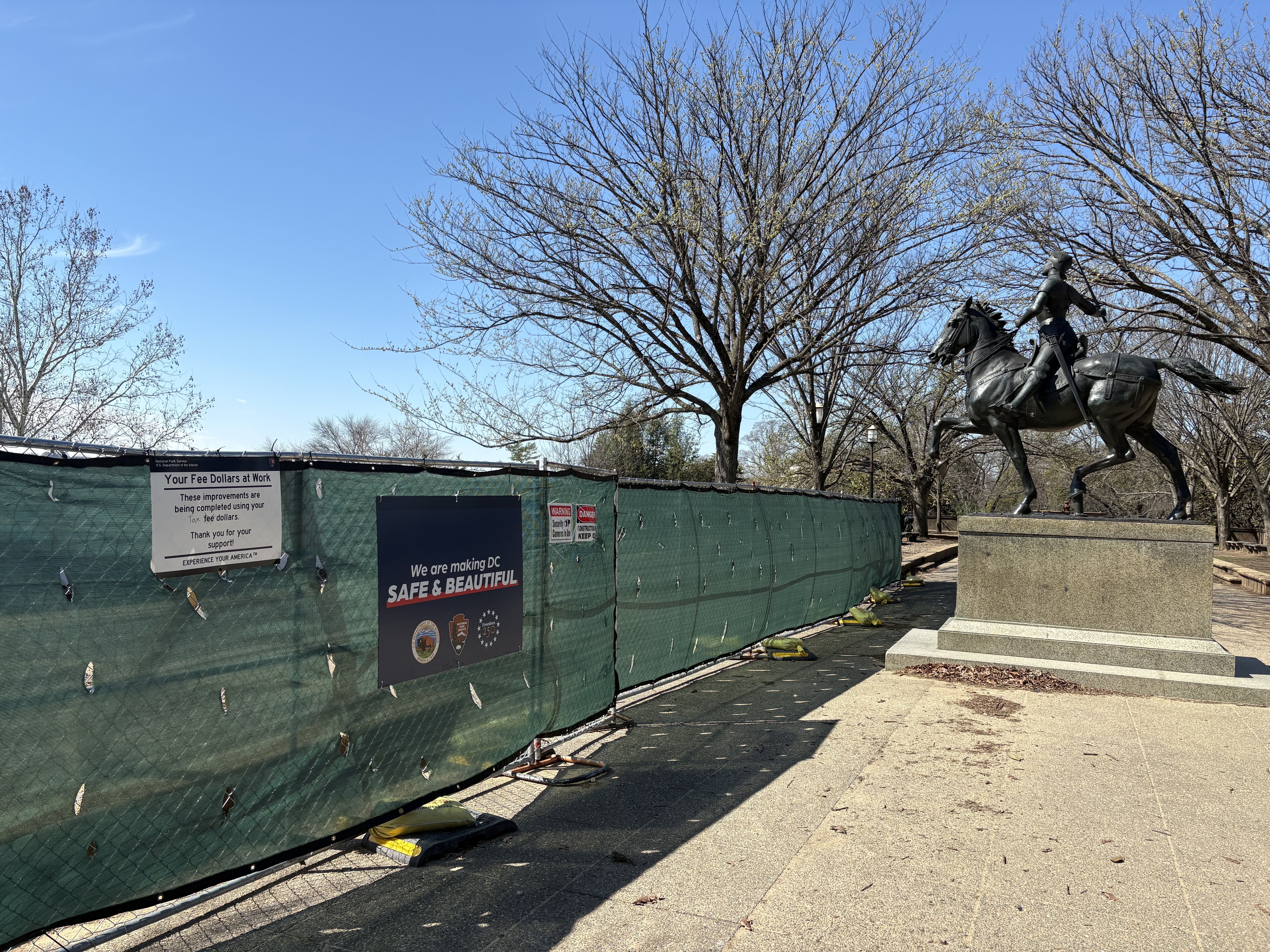 Bronze equestrian statue on a stone pedestal beside a green construction fence; leafless trees and a clear blue sky frame the scene, with a sign reading "We are making DC SAFE & BEAUTIFUL".