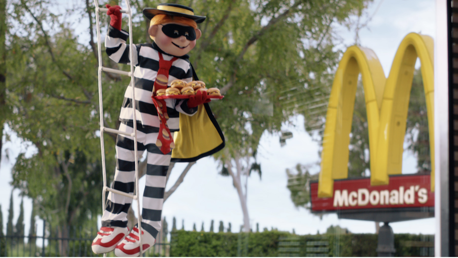 The Hamburgler stands on a rope ladder holding out with a tray of burgers in front of a McDonald's restaurant sign.