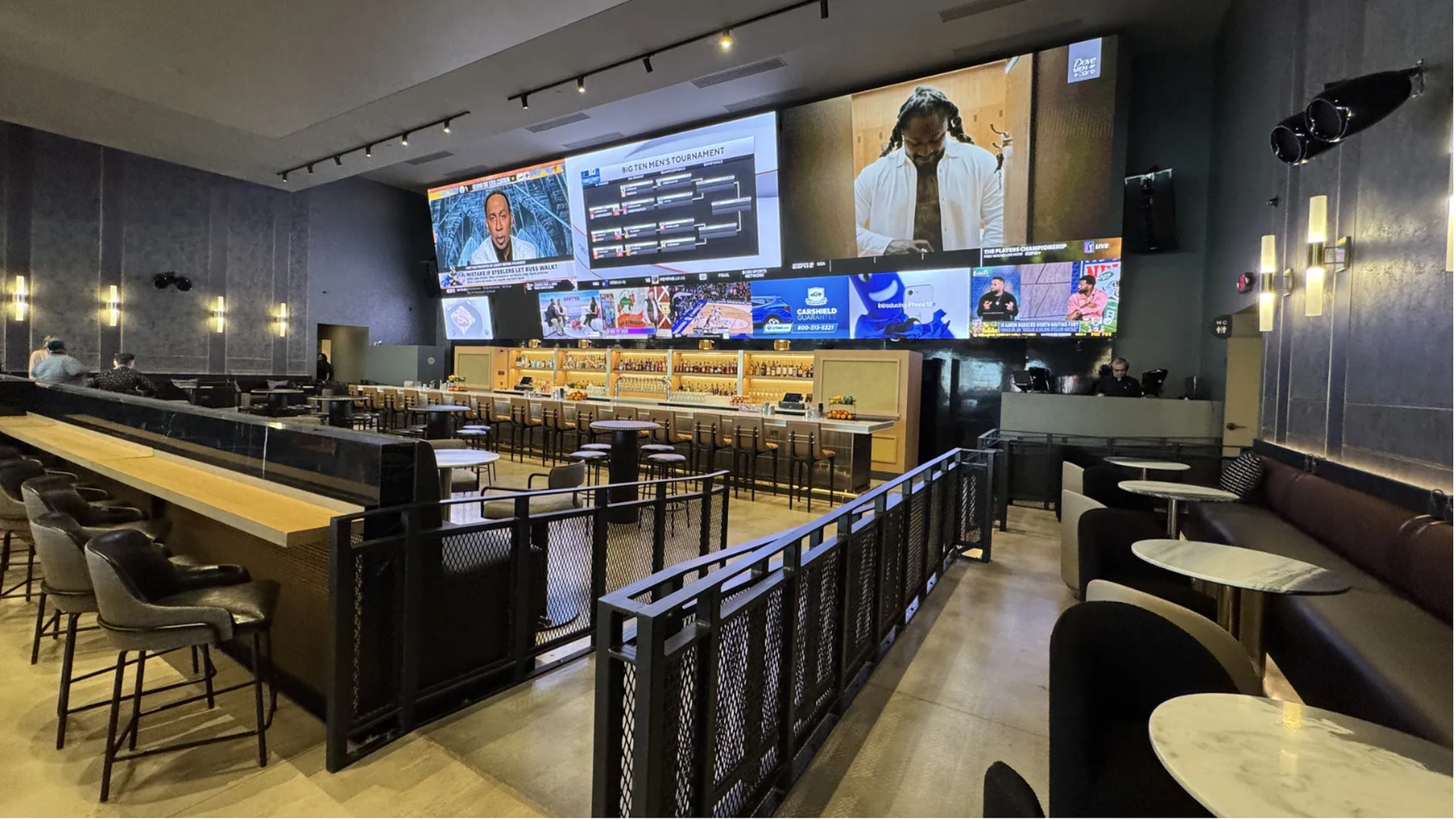 Modern sports bar with large video screens showing sports and news, black and brown seating, tall bar chairs, marble round tables, and warm overhead lighting on gray walls.