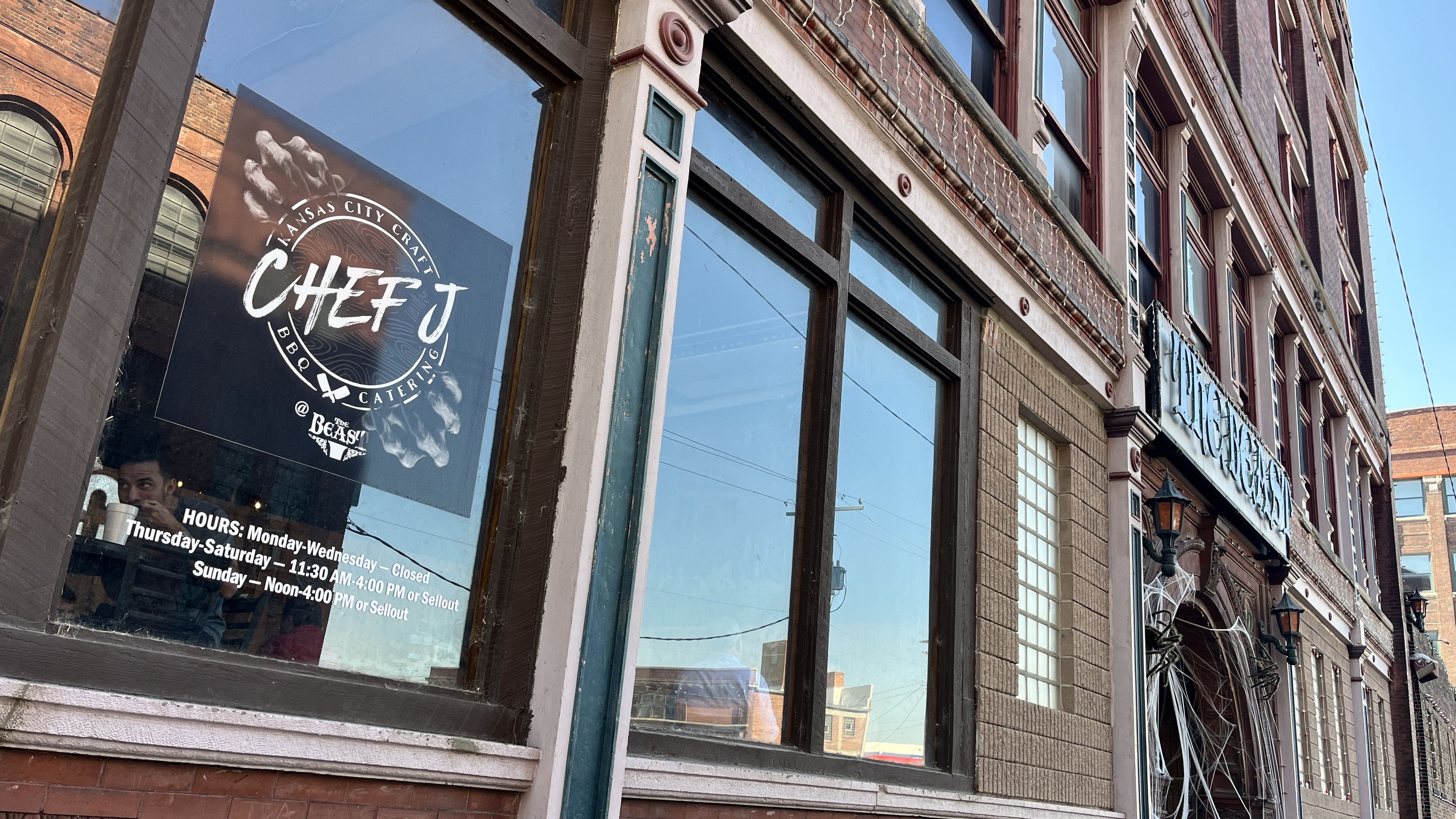 Window of Chef J Kansas City BBQ catering shows business hours; brick building with decorative details and a large sign reading "The Beast" above the door under clear blue sky.