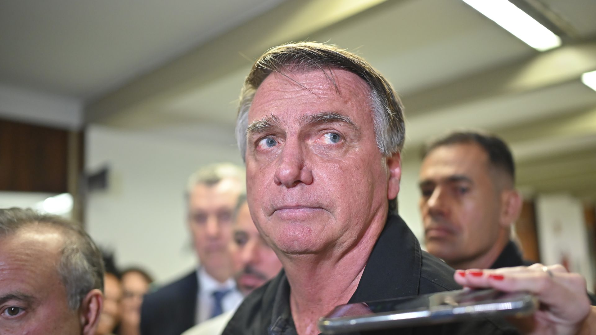  Former Brazilian President Jair Bolsonaro speaks to the press as a person holds a phone near his face.