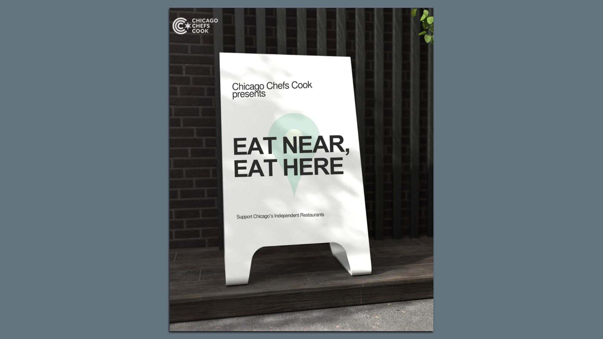 White sandwich board sign with black text reading "Chicago Chefs Cook presents EAT NEAR, EAT HERE" and "Support Chicago's Independent Restaurants" against a dark brick wall background.