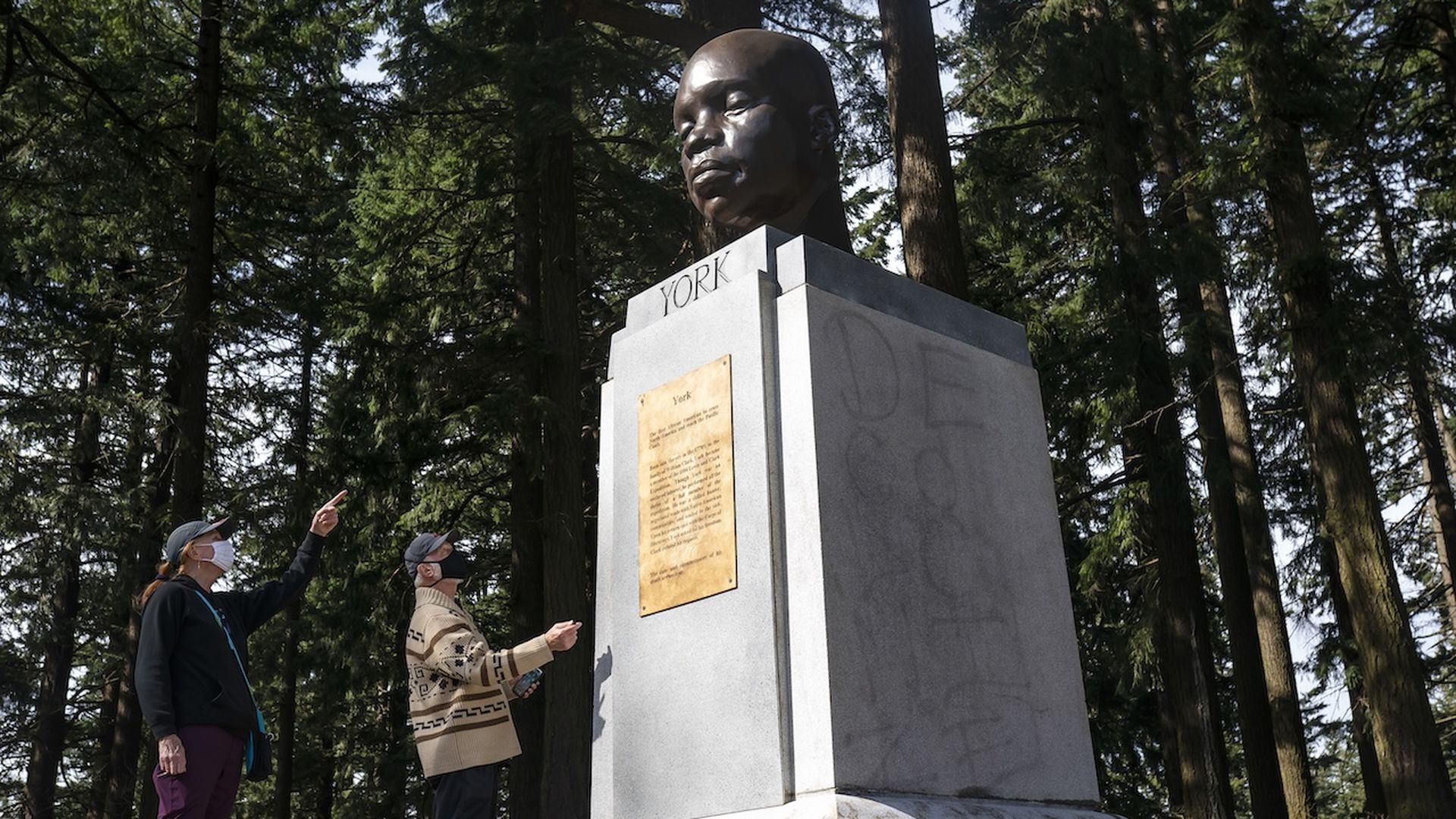 Two people wearing masks observe and point at a large monument of York, an African American member of the Lewis and Clark Expedition, set in a wooded area. The pedestal has a plaque and visible graffiti.