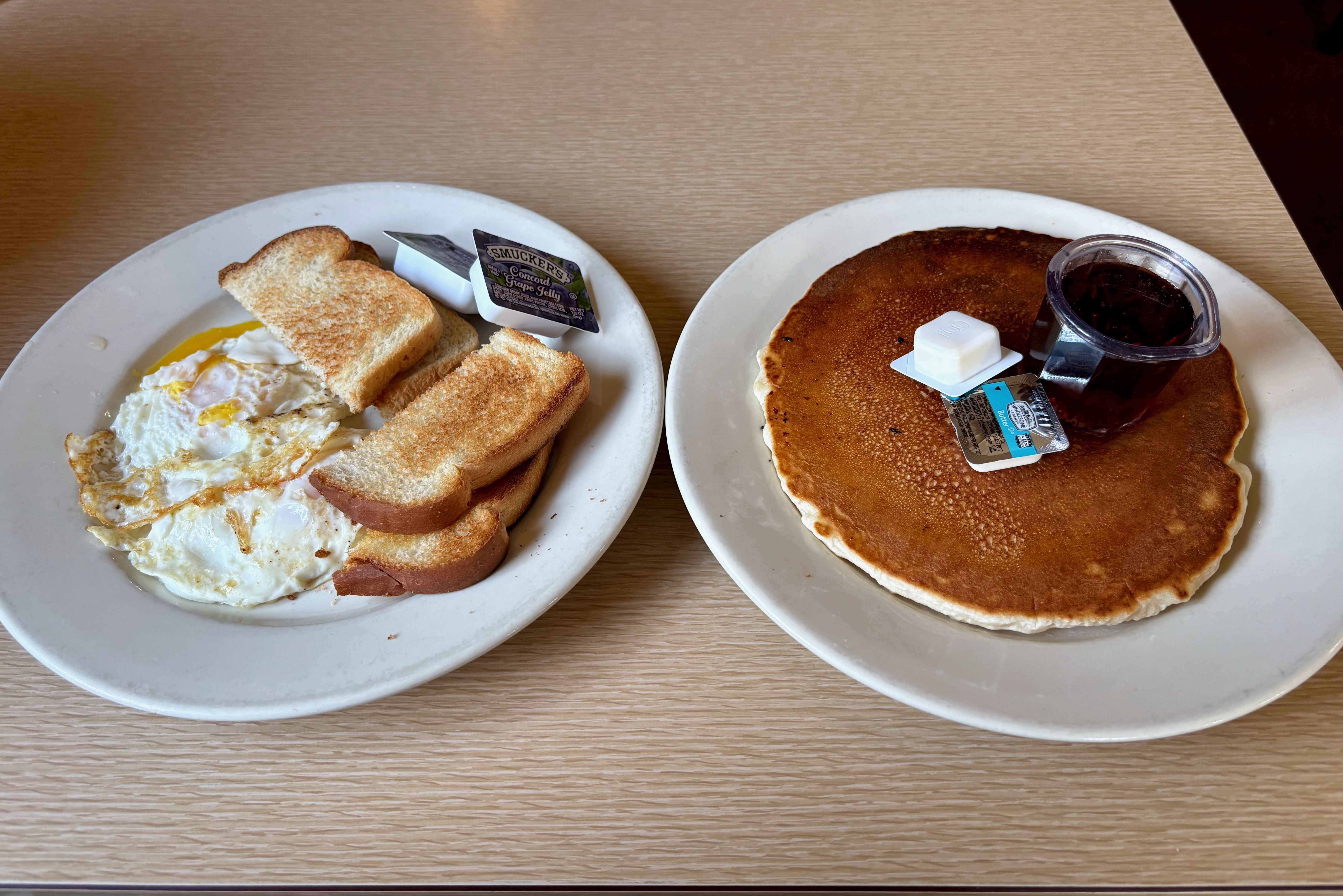 Two white plates on a light wood table: left plate has fried eggs with toast and a Smucker's Concord Grape Jelly packet; right plate holds two pancakes with butter and syrup cup.