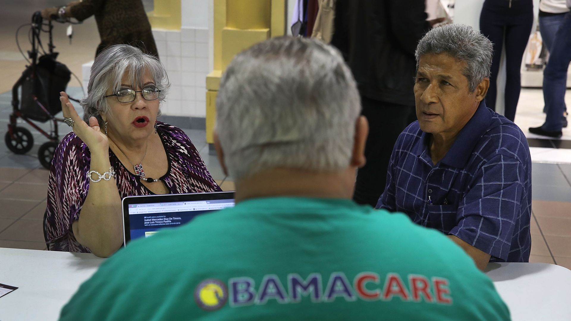 A man in a green shirt with the words "Obamacare" on the back helps two older adults sign up for health insurance.