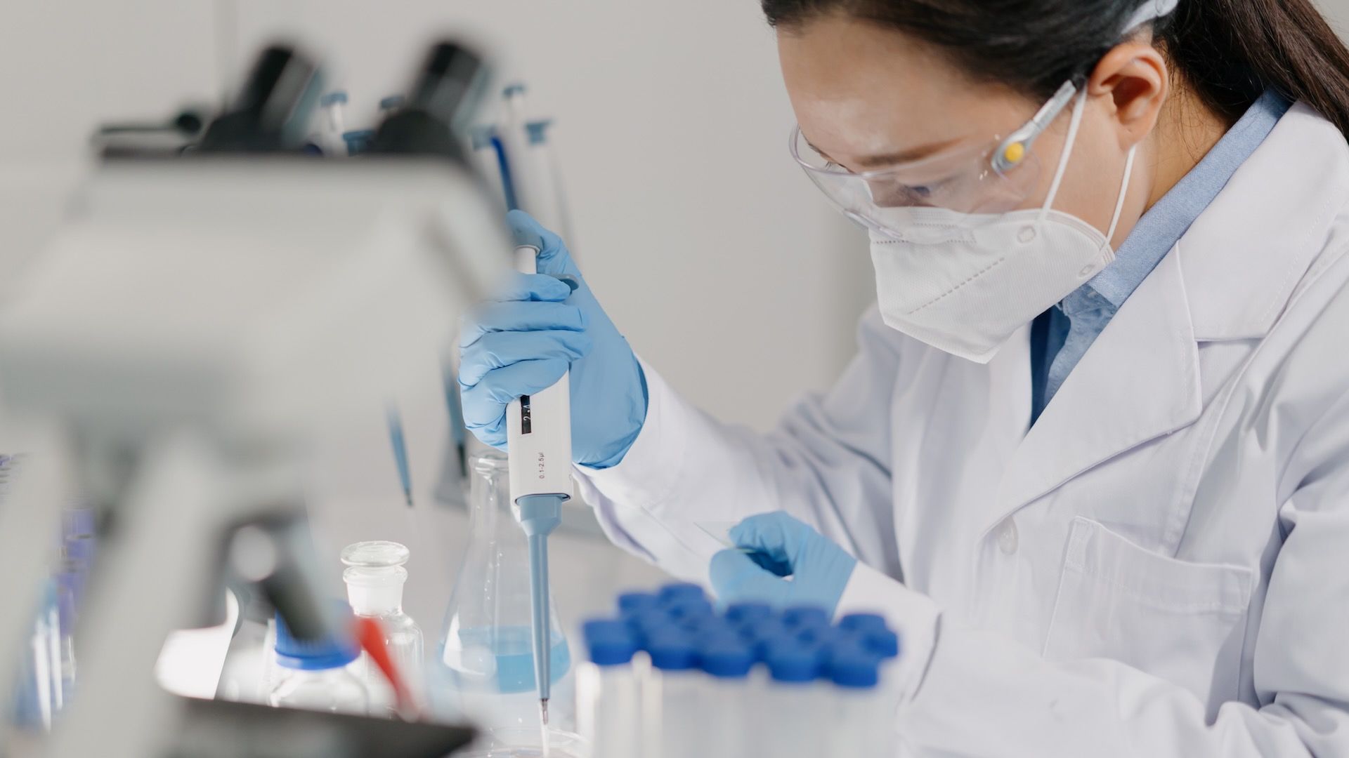A scientist in a white lab coat, mask, and safety goggles wears blue gloves and uses a pipette to transfer liquid into a flask in a bright laboratory with blurred equipment in the background.