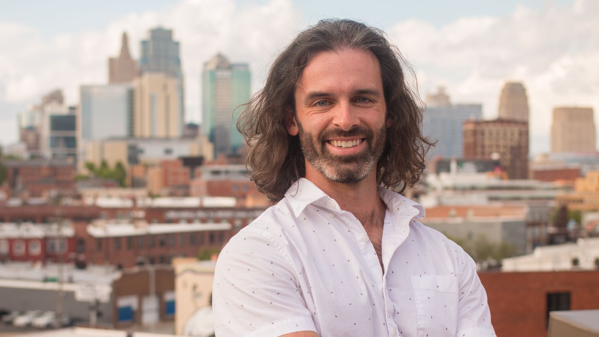 Smiling man with long brown hair and beard, wearing a white short-sleeve shirt with small patterns, stands with arms crossed on a rooftop with a city skyline in the background.
