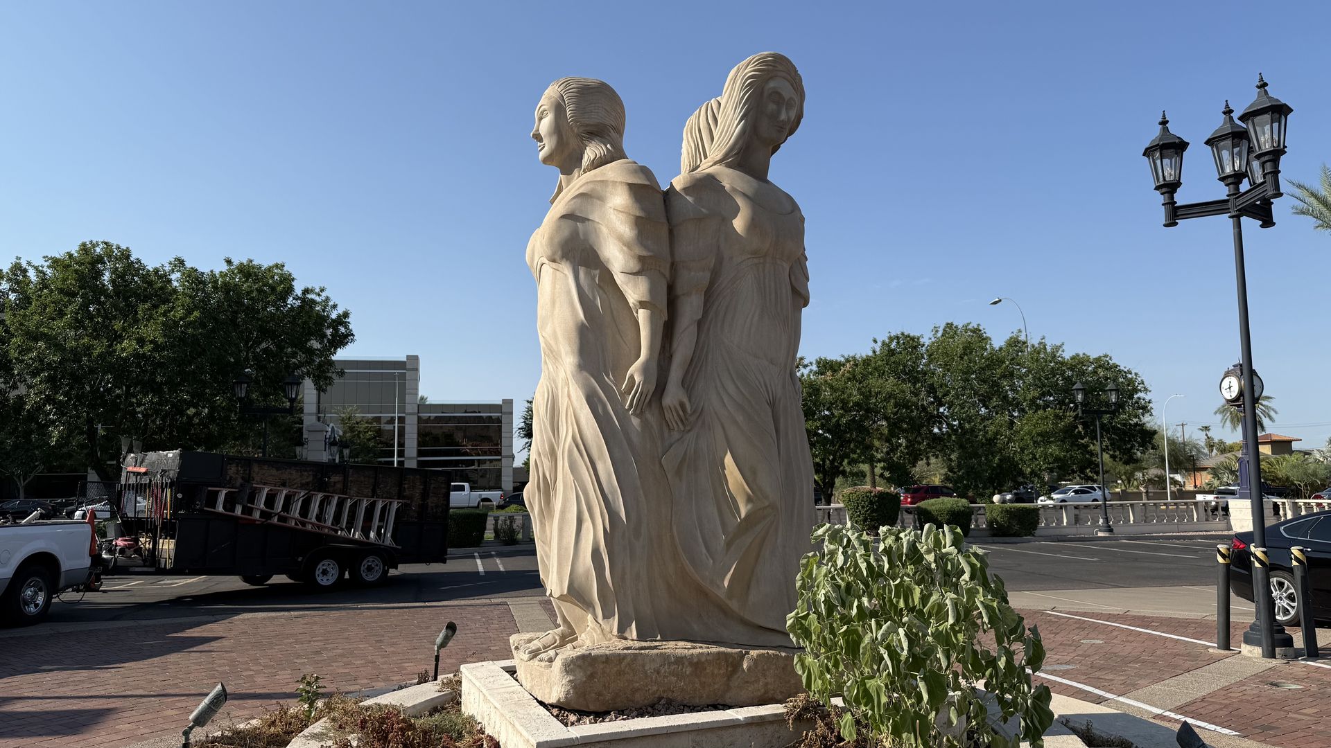 Beige stone sculpture of three women in flowing dresses with their backs to each other, standing in a circle. 