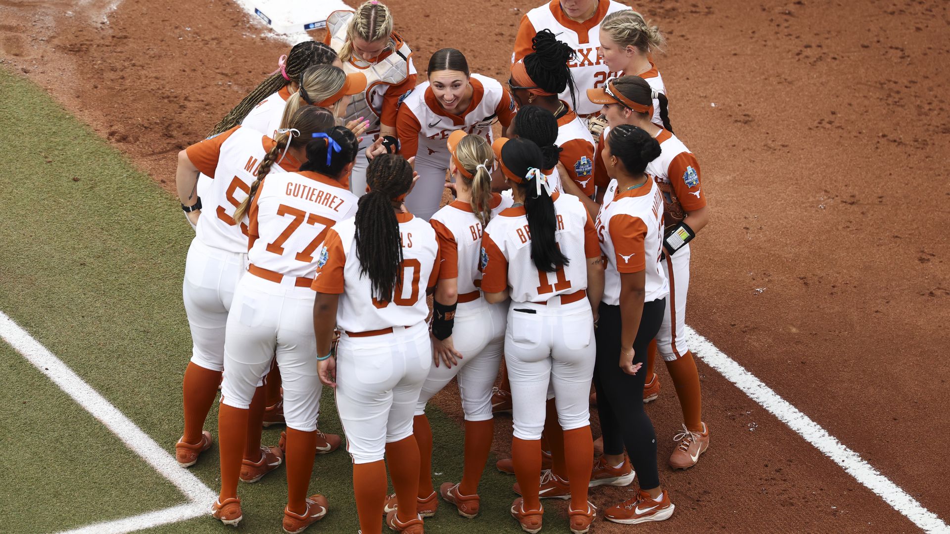 Softball team in white and orange uniforms huddles on field dirt near base, showing teamwork and focus during game.