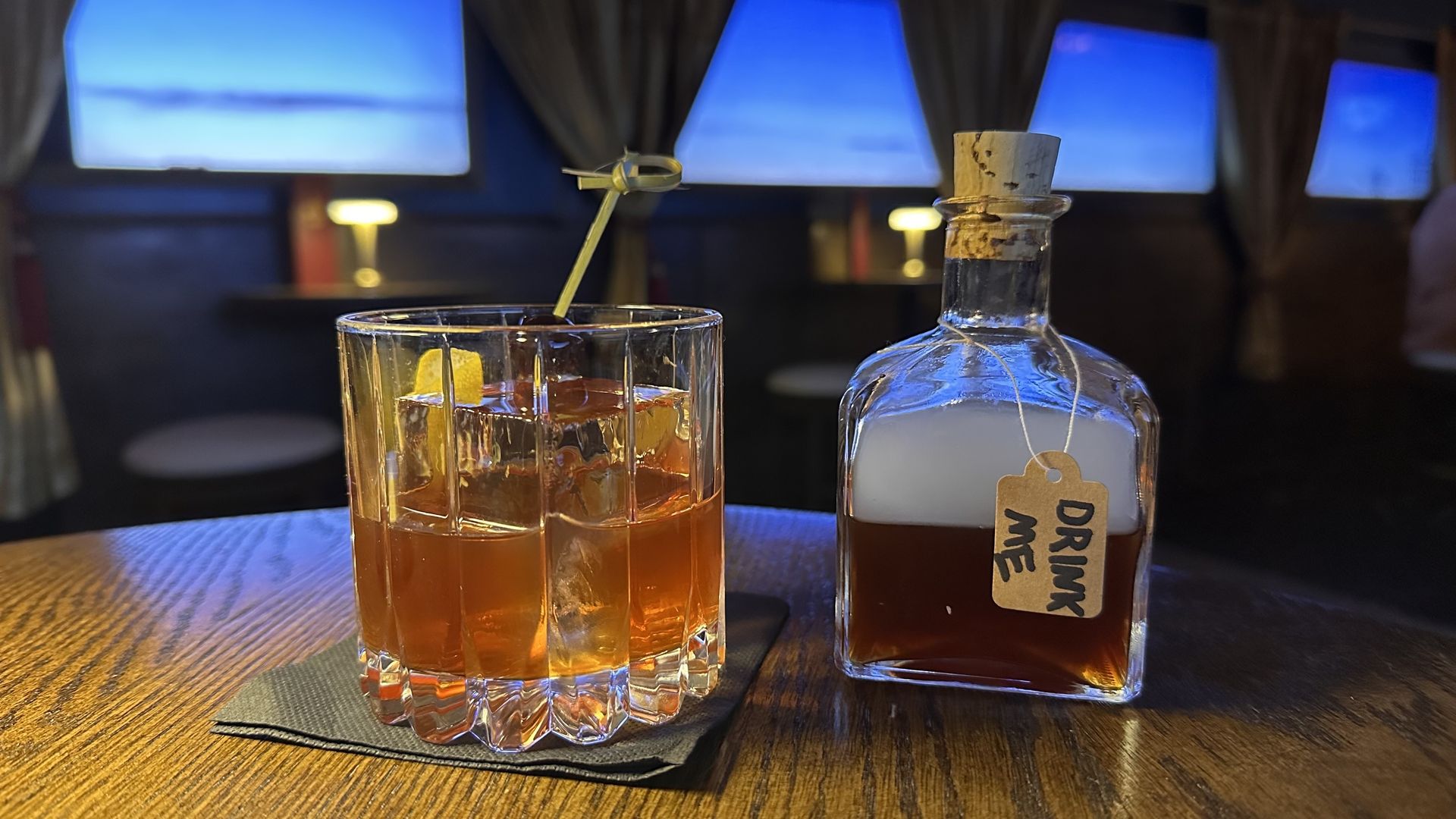 Glass of amber cocktail with ice and garnish on a wooden table beside a small bottle with a cork and tag reading "DRINK ME" in a dimly lit room with blue windows.